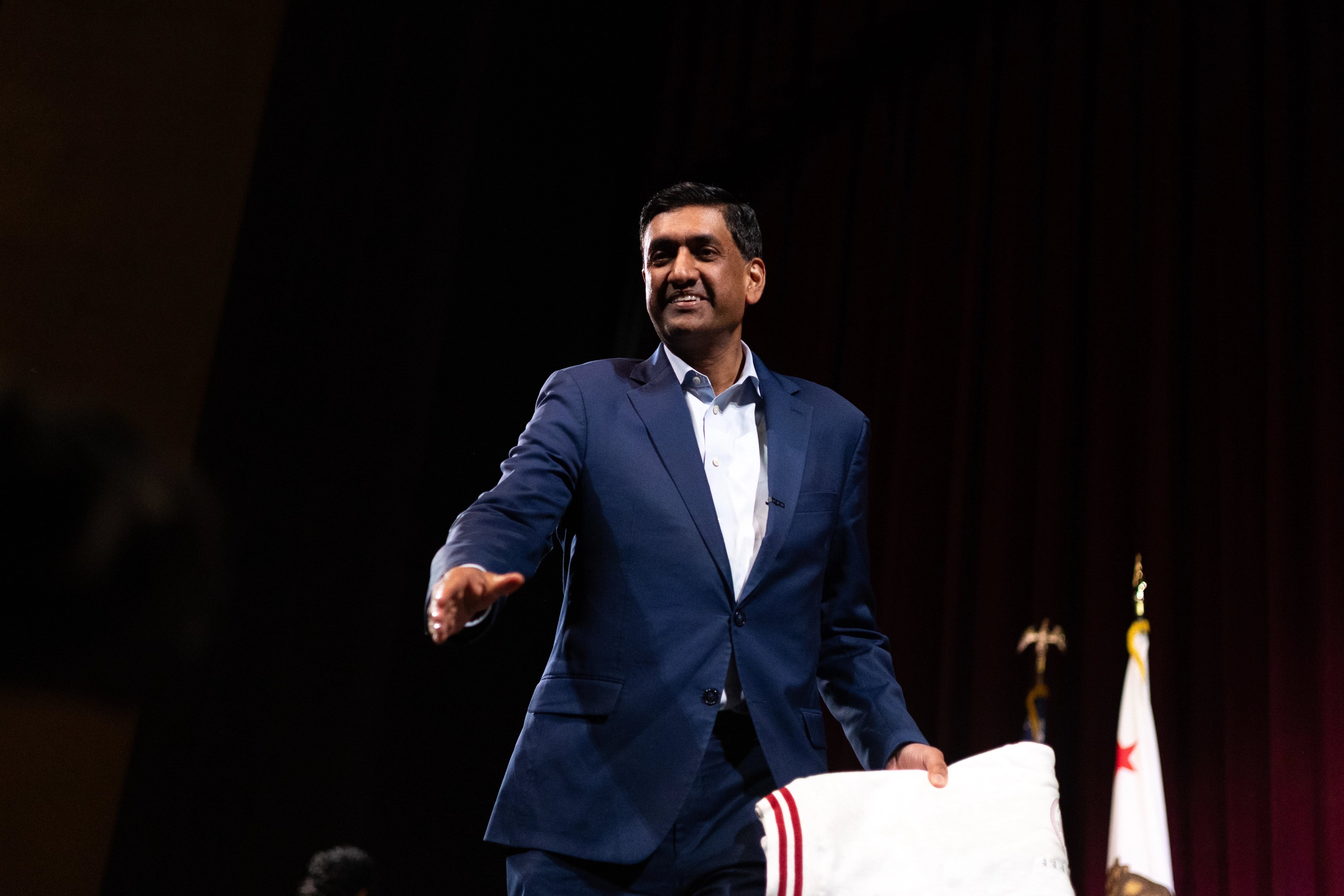 Ro Khanna in profile speaking into a microphone, his face partially lit by the bright white panel of a press videographer's camera light in the foreground, the back of a journalist's blonde hair silhouetted between them.