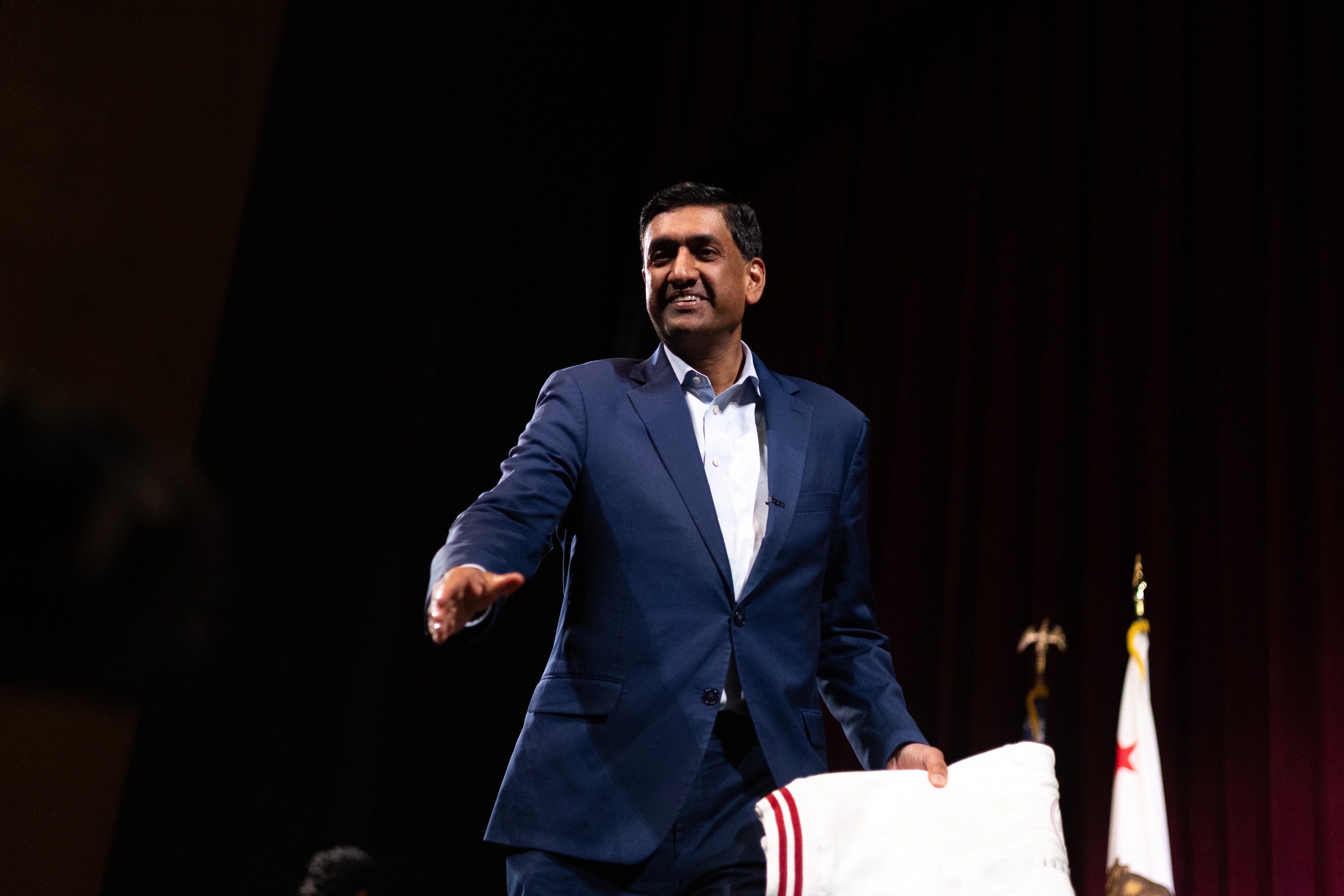 Ro Khanna in profile speaking into a microphone, his face partially lit by the bright white panel of a press videographer's camera light in the foreground, the back of a journalist's blonde hair silhouetted between them.