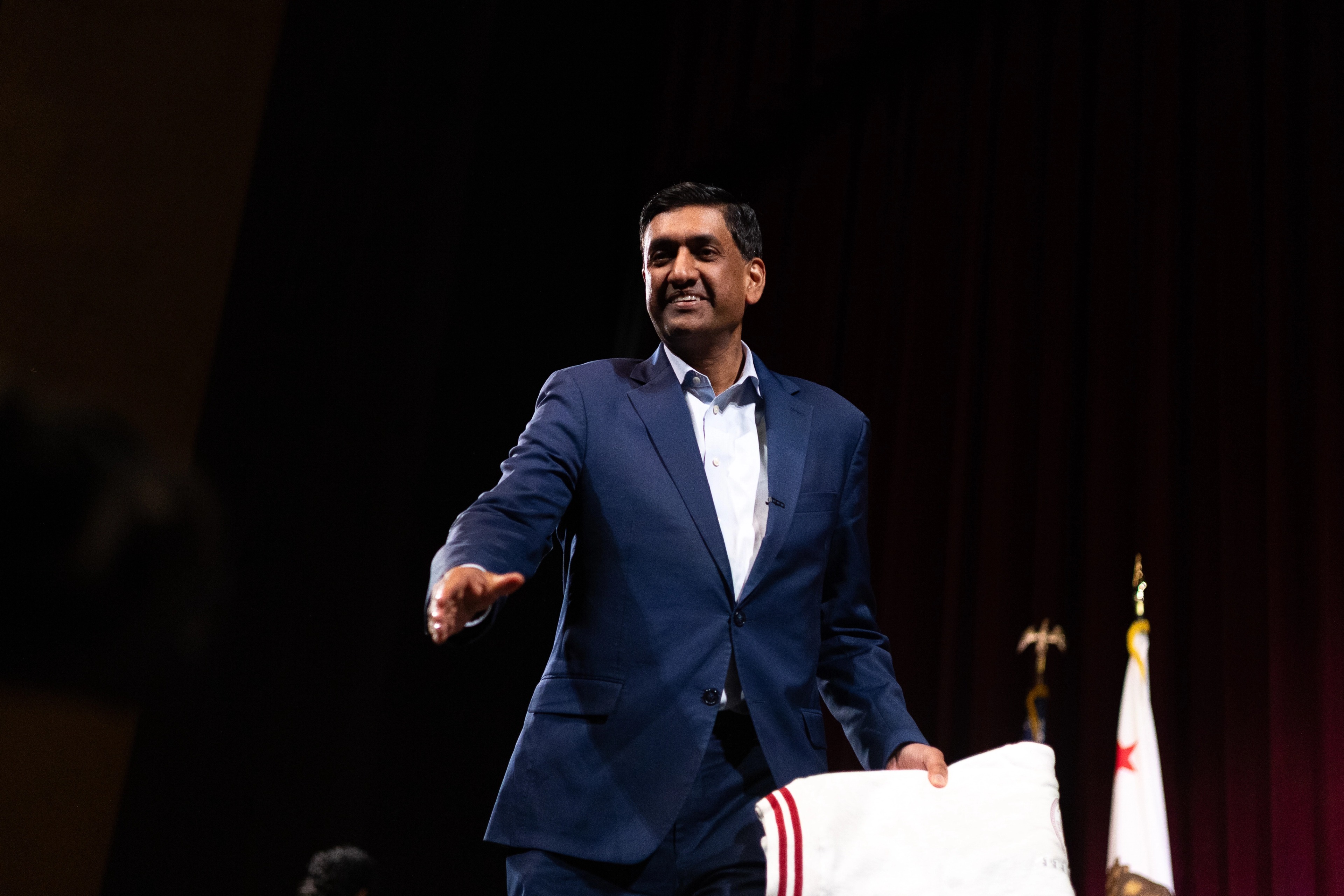 Ro Khanna in profile speaking into a microphone, his face partially lit by the bright white panel of a press videographer's camera light in the foreground, the back of a journalist's blonde hair silhouetted between them.