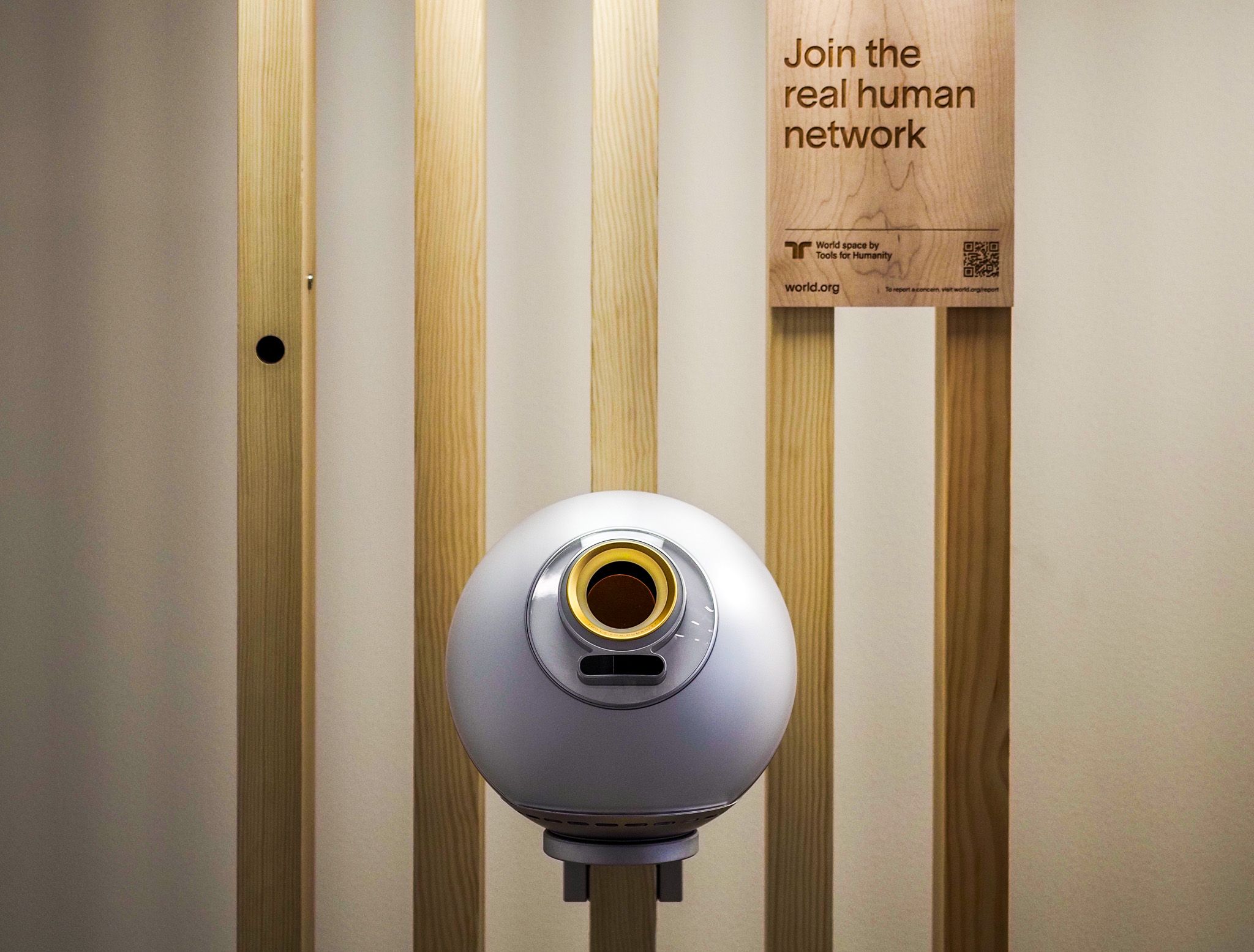 A spherical silver biometric scanning device with a single illuminated lens stands in front of vertical wood slats. A wooden sign above it reads 'Join the real human network' with the word 'world' and a circular logo.