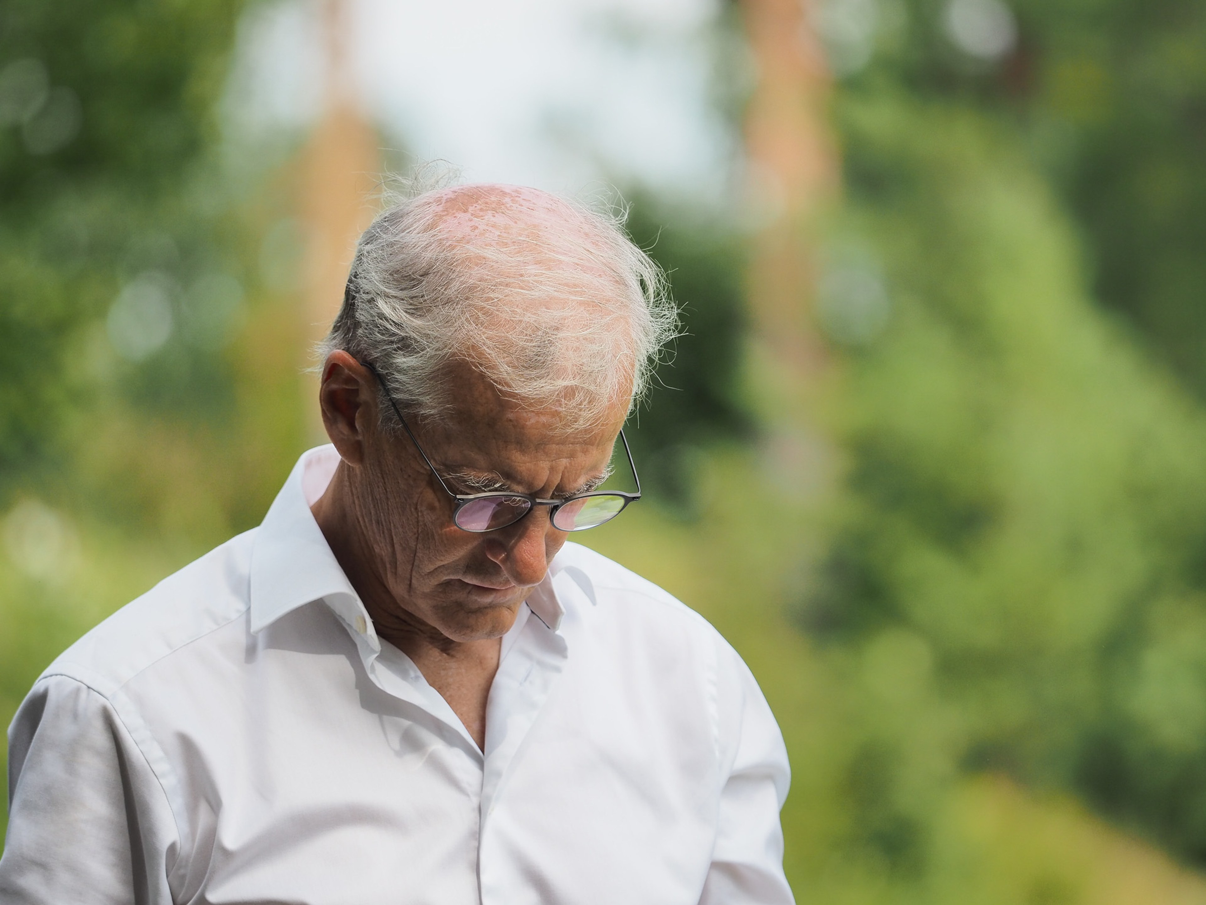 Three-quarter portrait of Jonas Gahr Støre with his head bowed, eyes down, wearing thin-framed glasses and a white open-collar shirt, against a softly out-of-focus background of summer foliage.