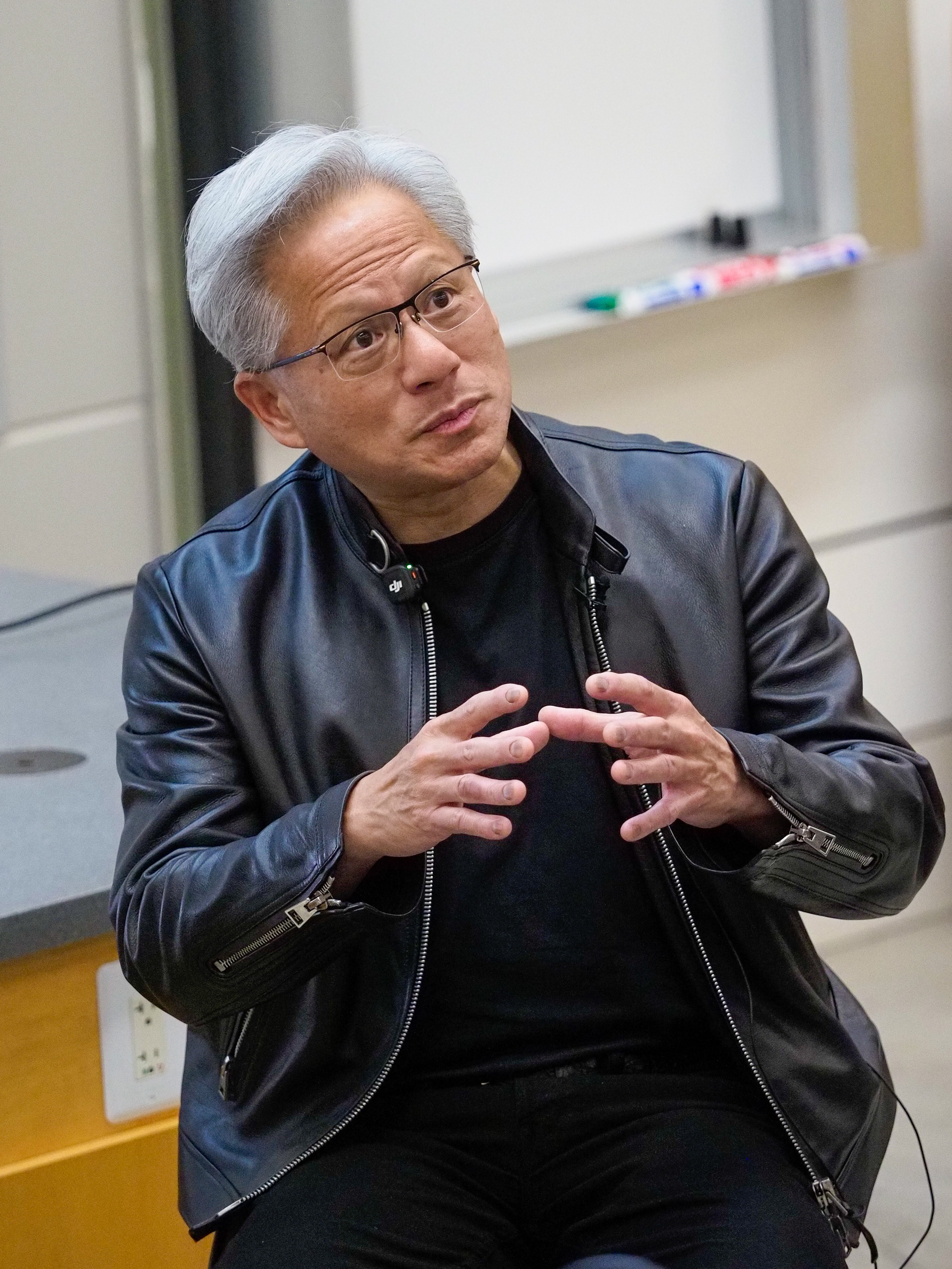 NVIDIA CEO Jensen Huang in a black leather jacket and round glasses, mid-gesture with both hands raised, photographed in profile against a whiteboard with markers visible on the ledge.