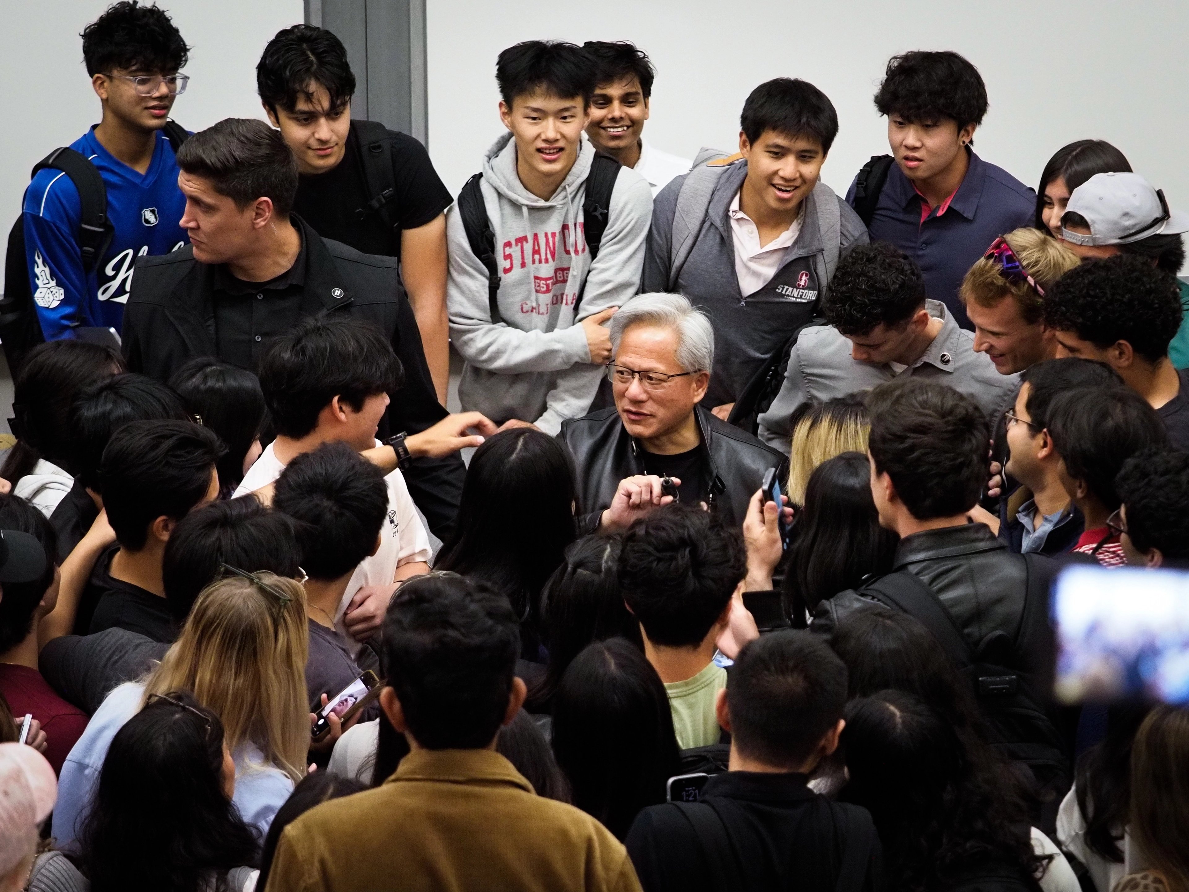 Jensen Huang at the center of a tight crowd of students; a young man with curly hair leans in for a selfie at left holding up his phone, another student in a white shirt smiles between them.