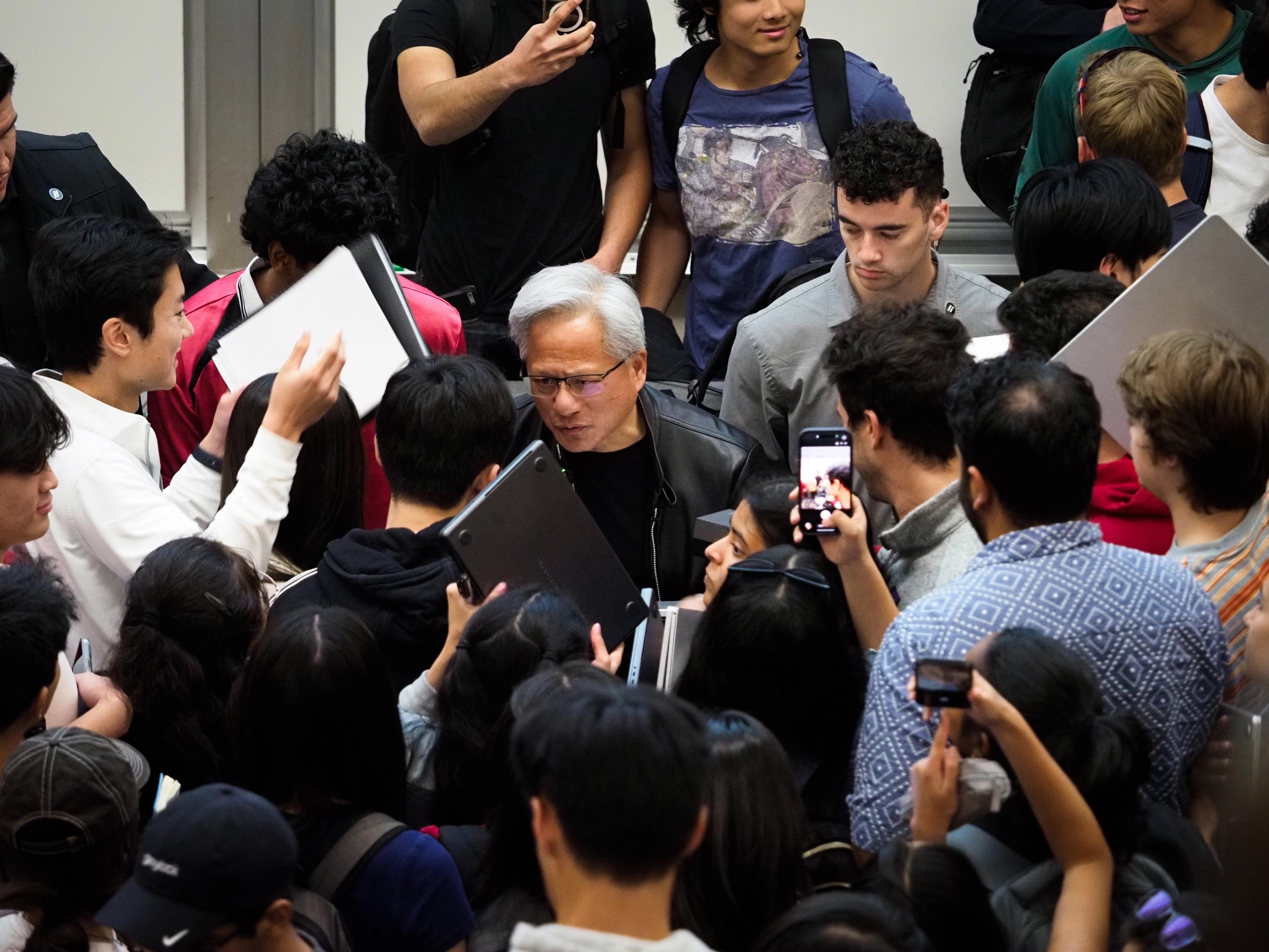 Jensen Huang at the center of a packed crowd of Stanford students, several students standing tall behind him with phones and a backpack visible, a woman in the foreground holding up a phone toward the scene.