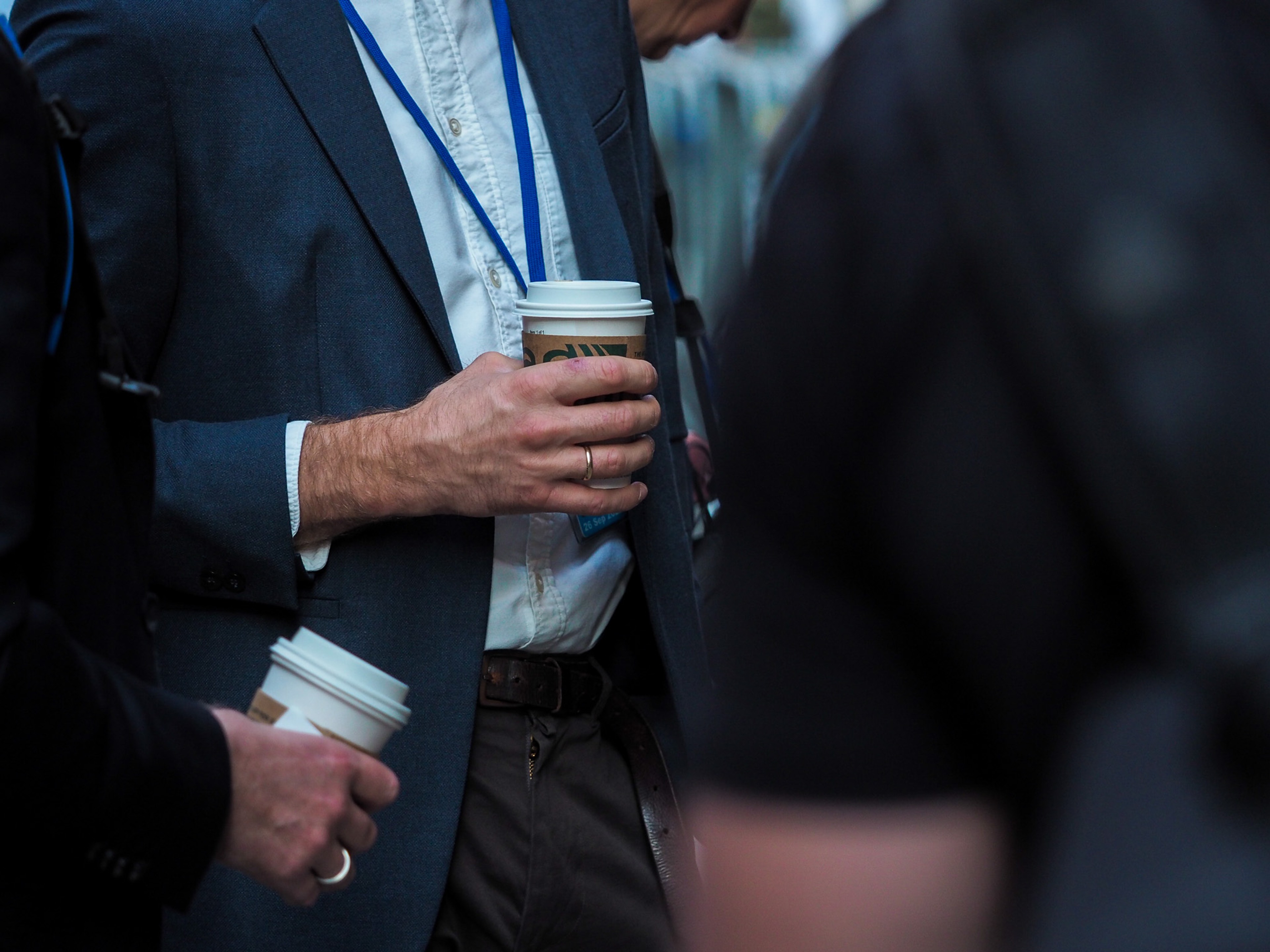 Tight torso shot of two men in dark suits holding takeaway coffee cups, blue United Nations lanyards around their necks, faces out of frame.