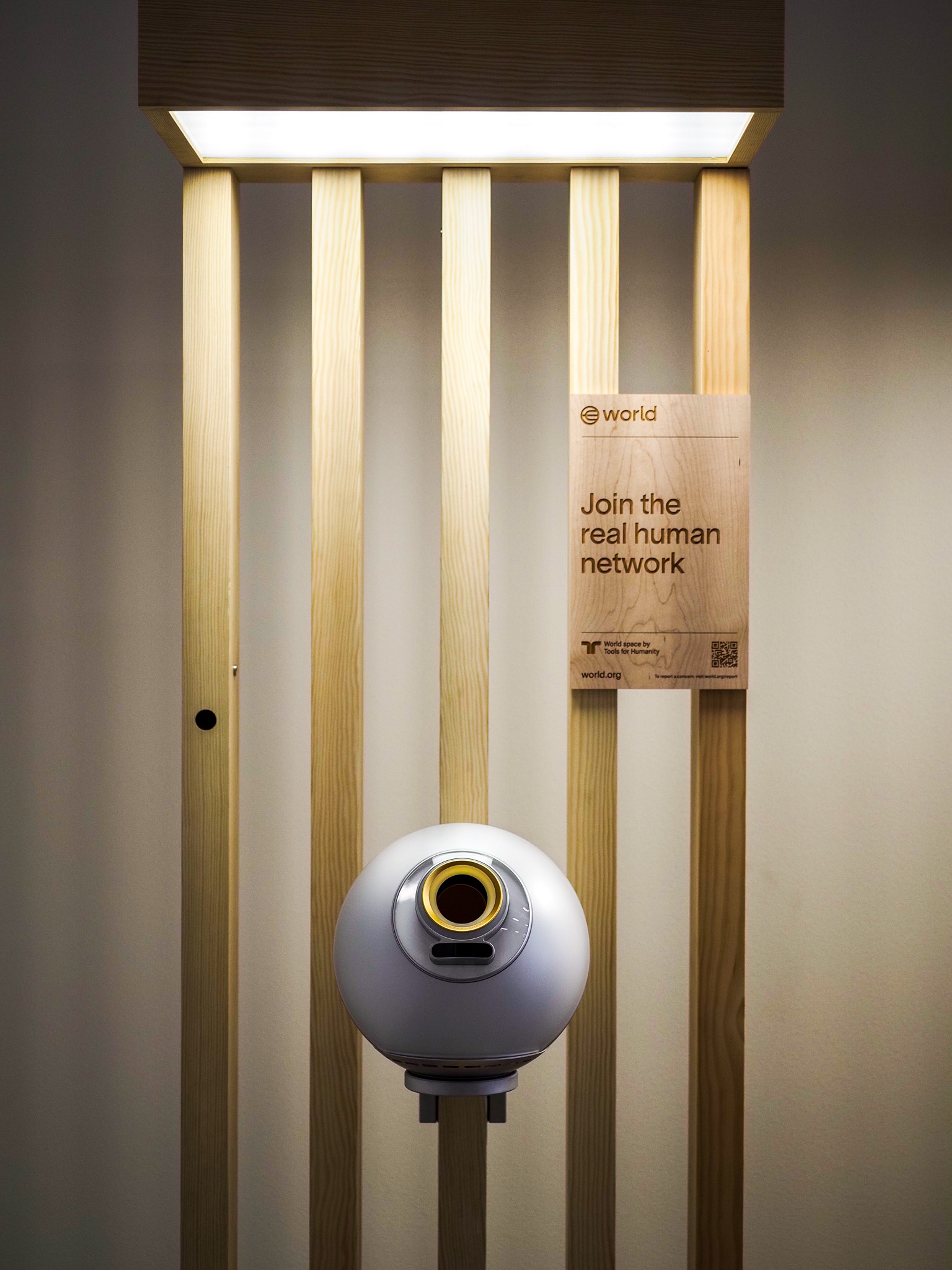 Wide view of a Worldcoin Orb biometric scanner mounted in a slatted-wood enclosure with overhead lighting. A wooden sign reads 'Join the real human network'.
