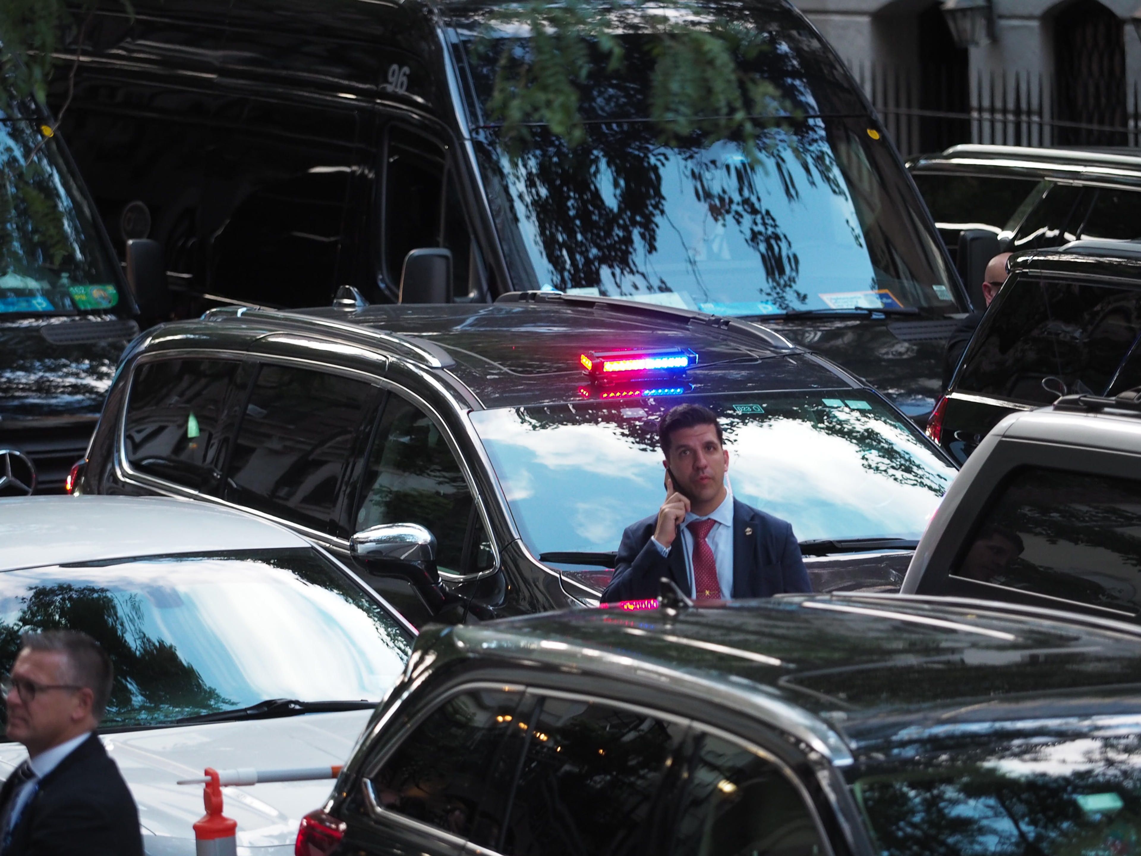 A man in a dark suit and red tie takes a phone call standing between two black SUVs of a motorcade, a red and blue police light bar visible above the cars.