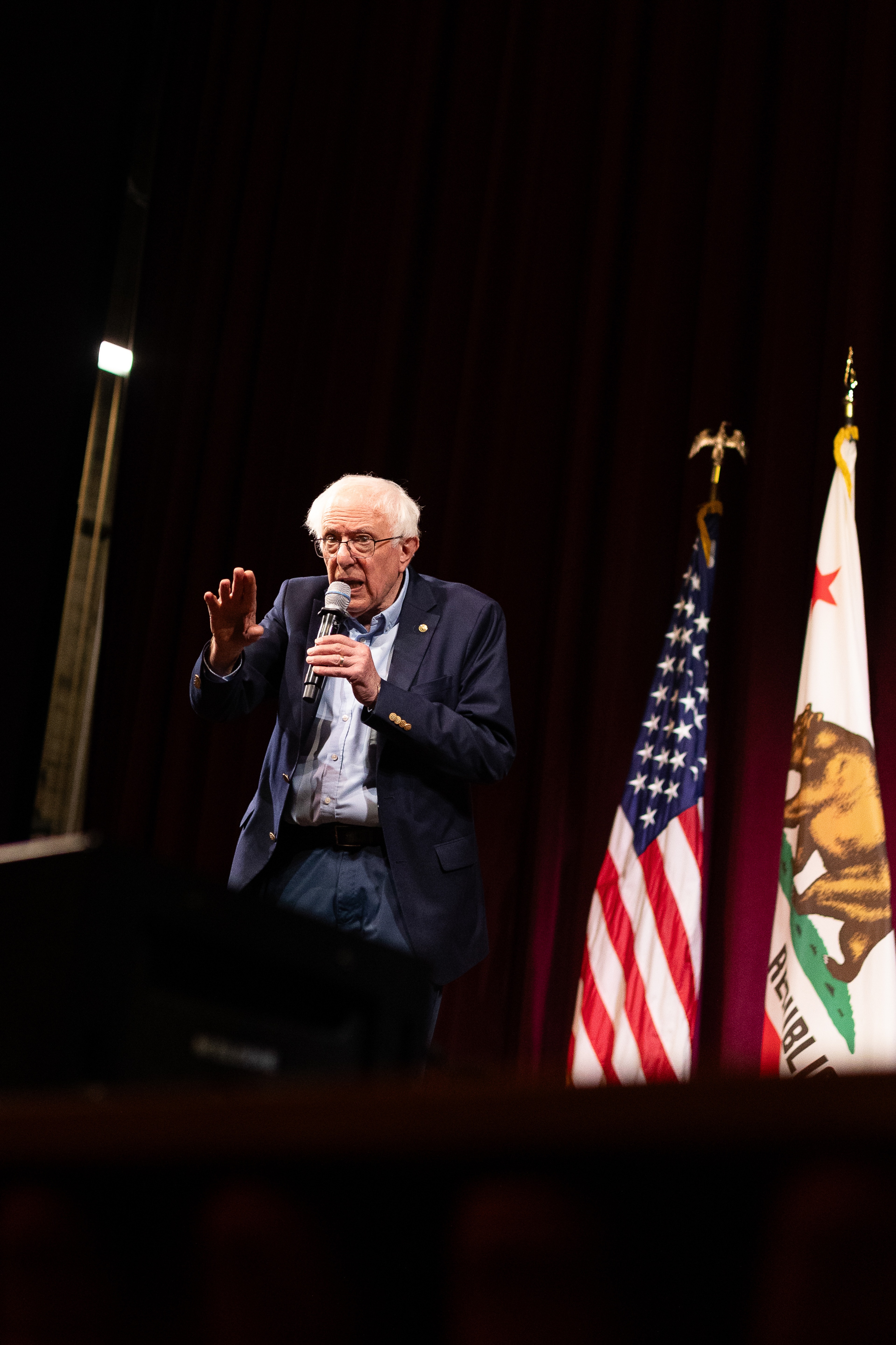 Tight three-quarter portrait of Bernie Sanders speaking into a microphone, partial American flag visible to the right, dark stage background.