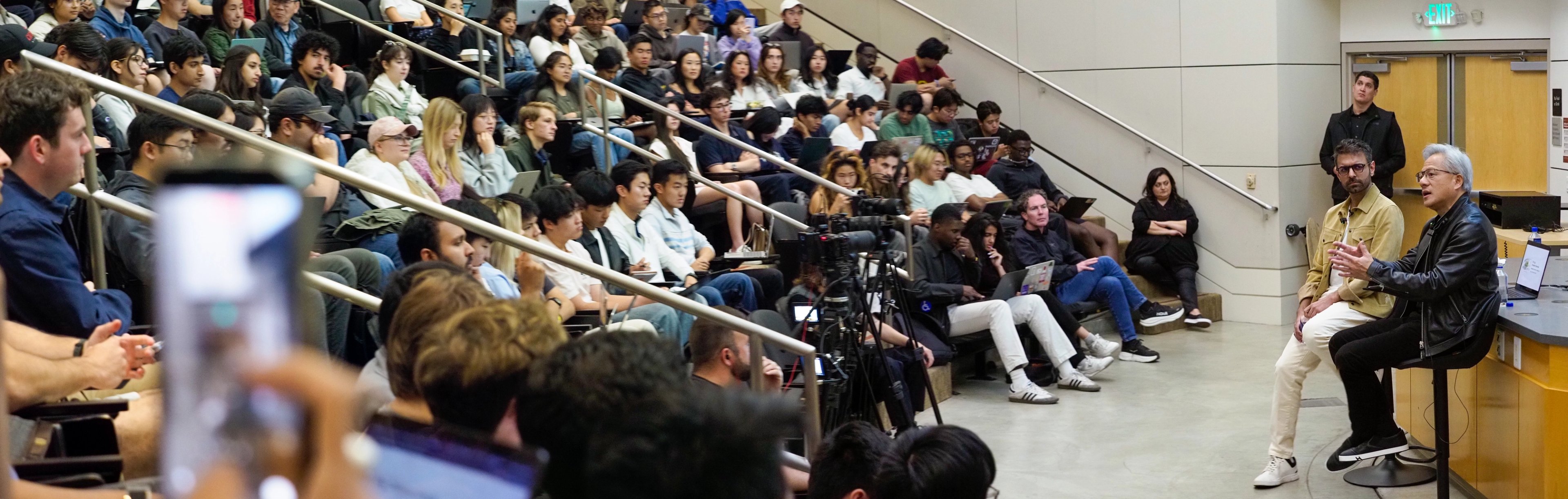 Wide panoramic view of a tiered Stanford lecture hall, students seated in rows watching Jensen Huang and Anjney Midha at the front, video cameras mounted on tripods between them.