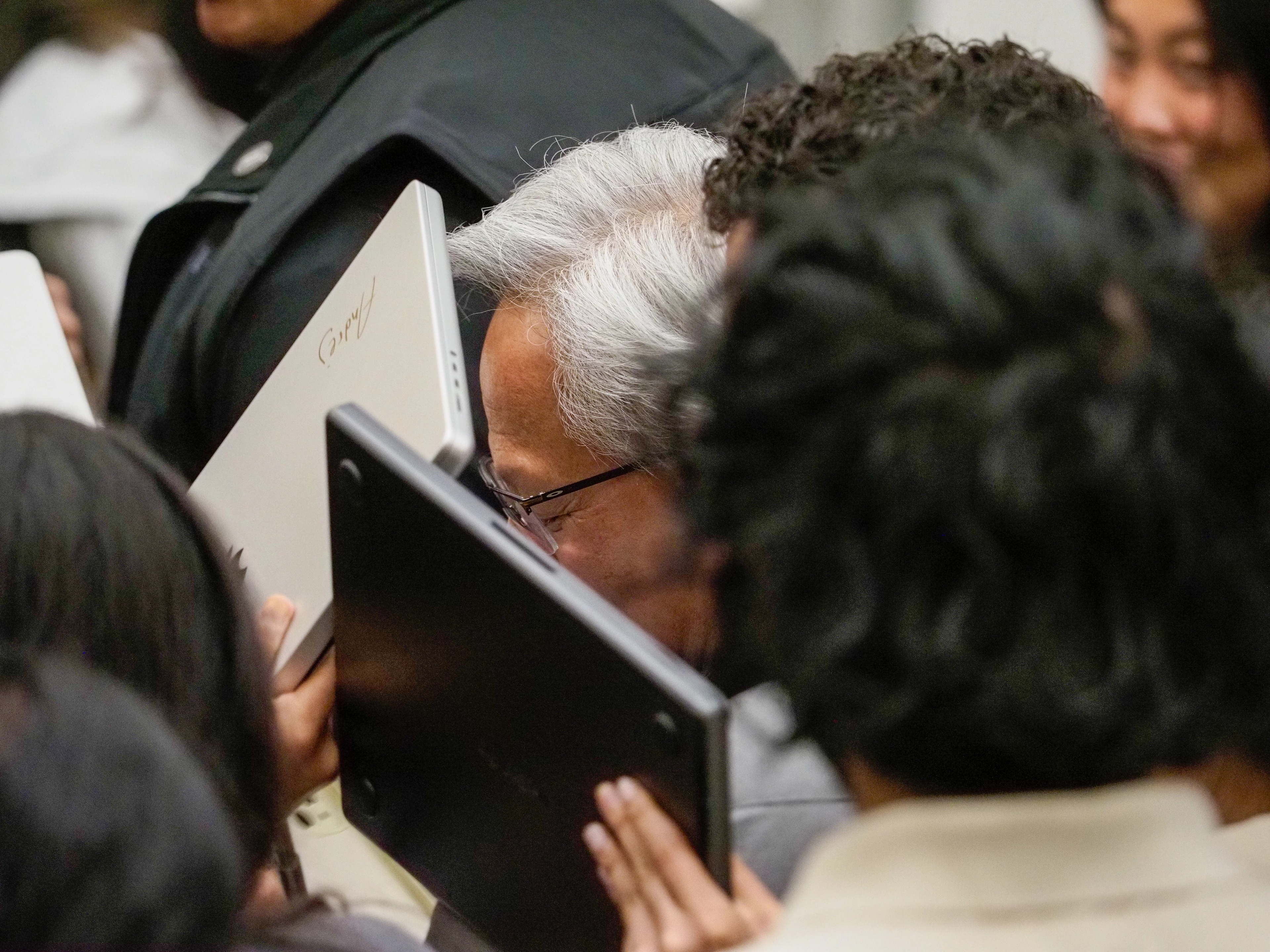 Jensen Huang's face mostly obscured behind two laptops held up in front of him by students, only his grey hair, glasses, and eyes visible between the screens.