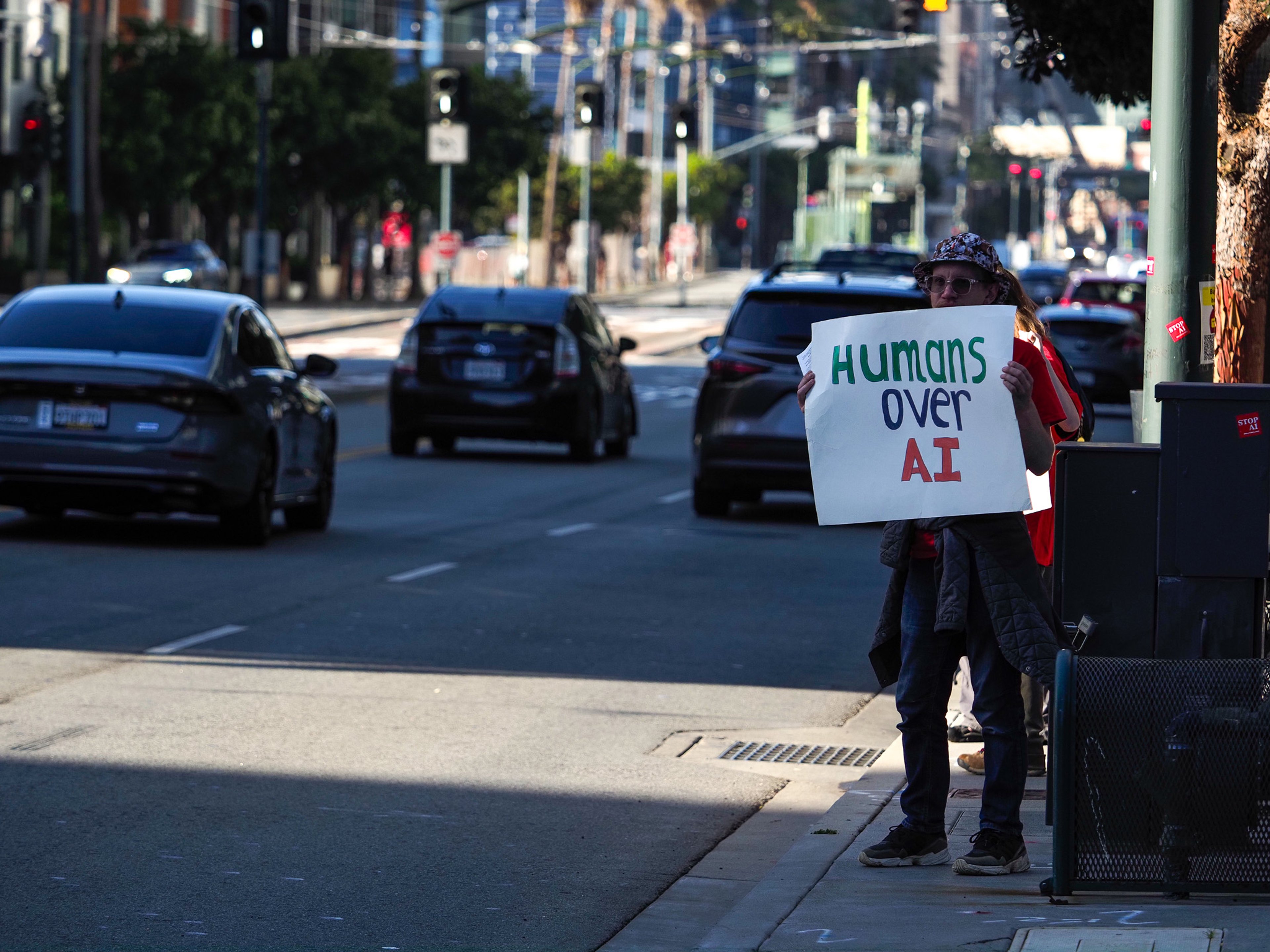 A lone protester in a bucket hat stands on a sidewalk holding a homemade sign reading 'Humans over AI' as cars pass on a wide downtown street.