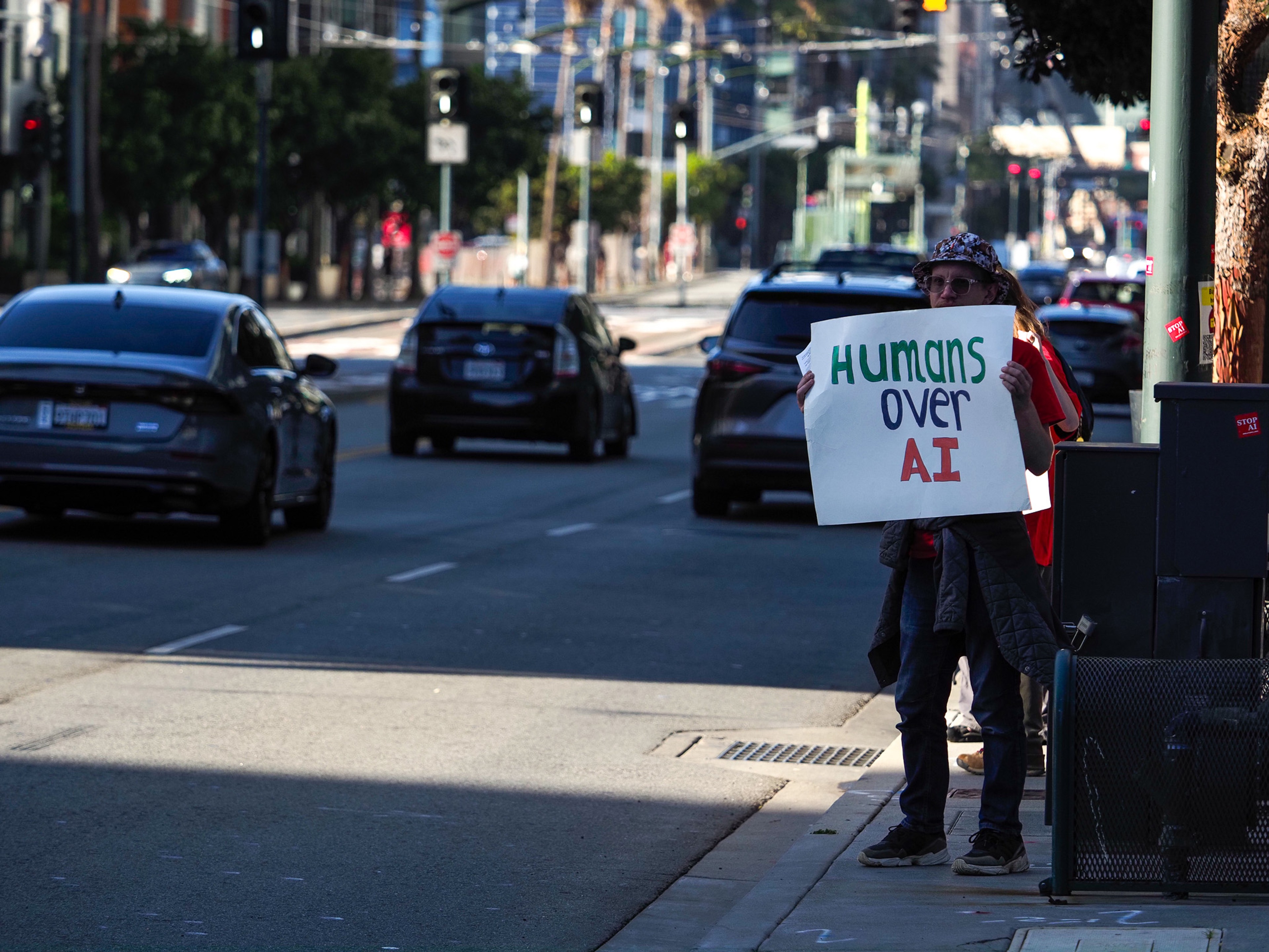 A lone protester in a bucket hat stands on a sidewalk holding a homemade sign reading 'Humans over AI' as cars pass on a wide downtown street.