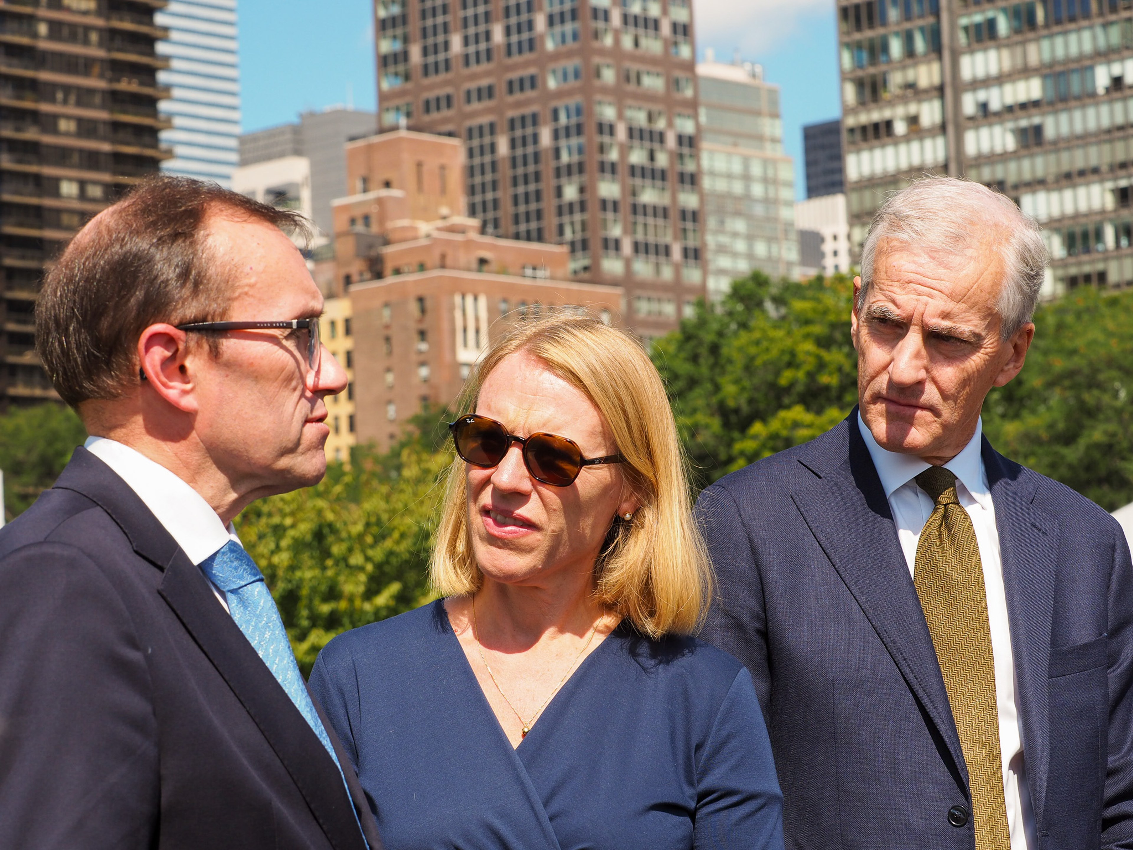 Three figures outdoors in front of the Manhattan skyline — a man in glasses in profile mid-speech (Espen Barth Eide), a blonde woman in sunglasses and a navy dress (Anniken Huitfeldt), and a gray-haired man in a navy suit and gold tie (Jonas Gahr Støre).