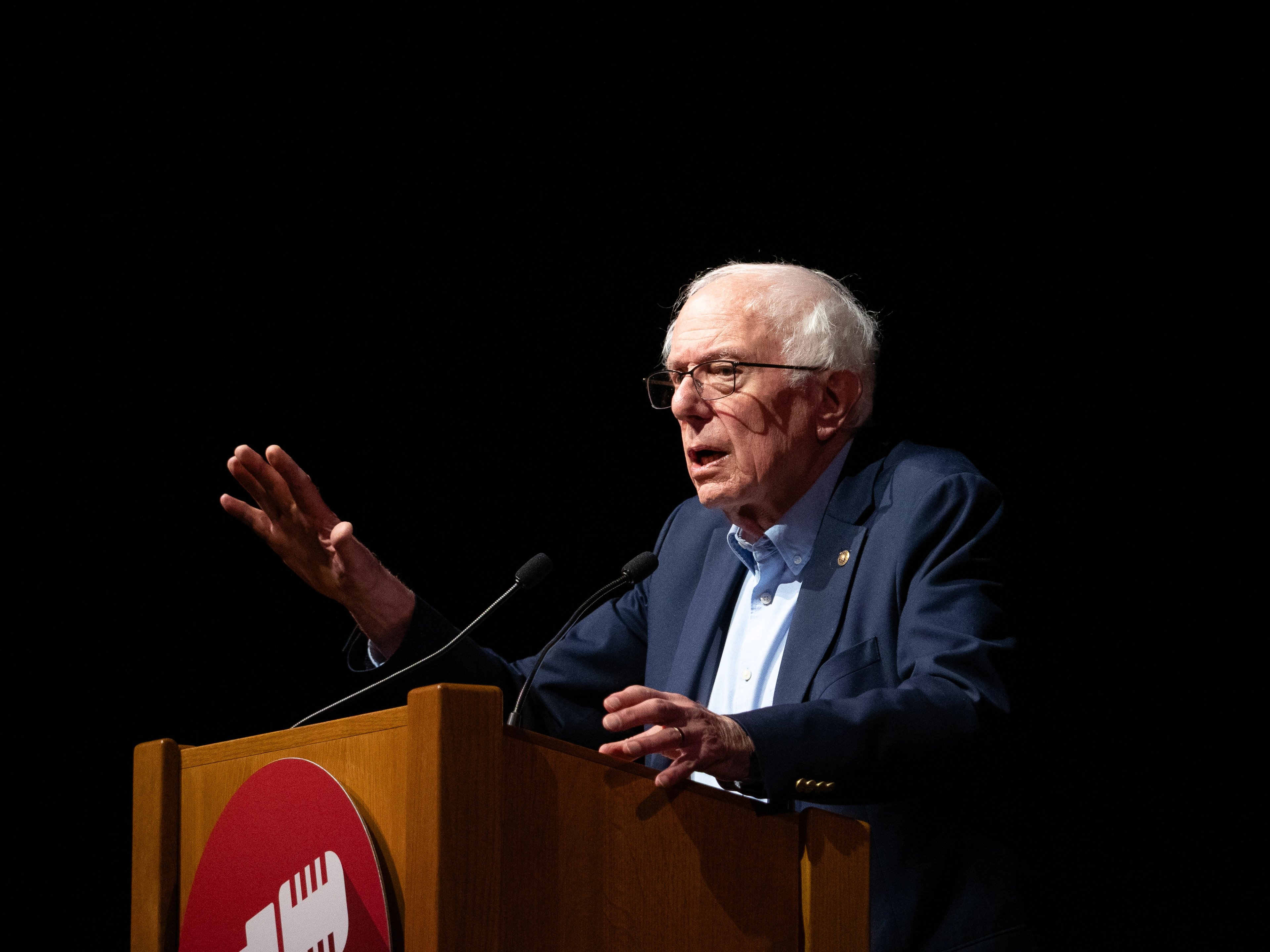 Bernie Sanders behind a wooden podium, both hands raised palm-out mid-gesture, the red microphone-icon logo on the front of the podium, microphones at chest height.