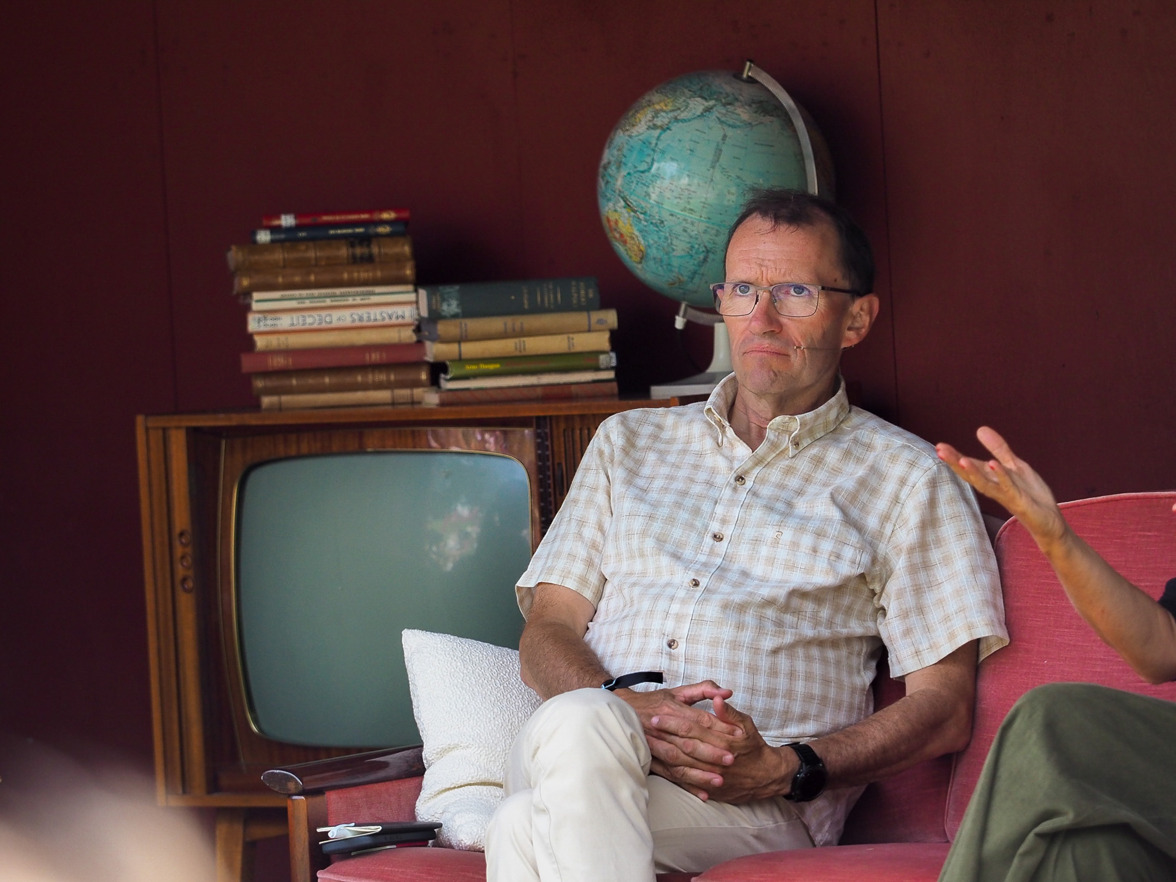 Espen Barth Eide seated in a coral-pink upholstered chair, hands clasped on his lap, wearing a beige checked short-sleeved shirt and thin-framed glasses, with a vintage television, a stack of old hardback books, and a globe arranged on a wooden cabinet behind him.