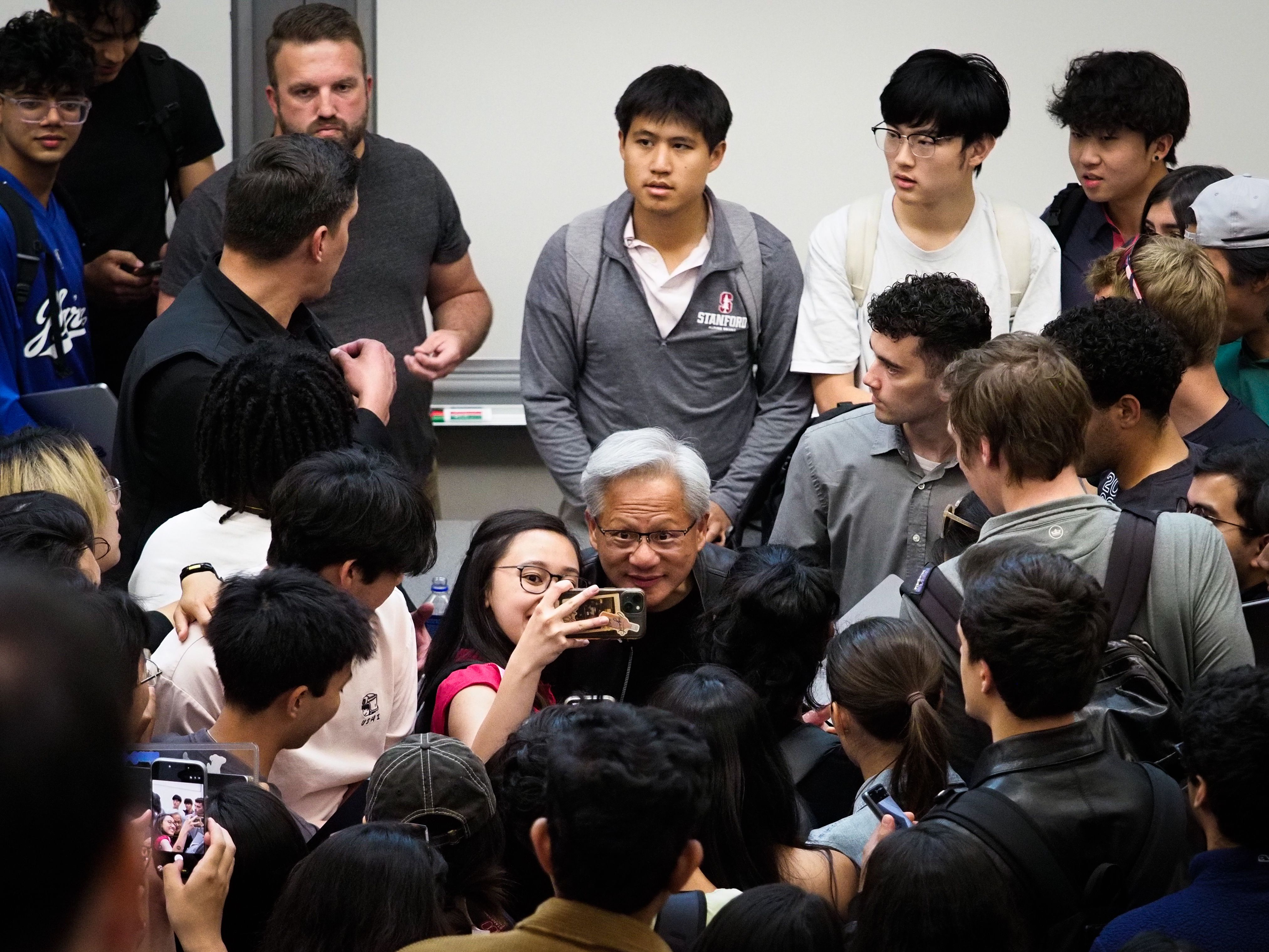 Jensen Huang at the center of a tight cluster of Stanford students, a young woman in a white shirt and red scarf in the foreground looking up at him smiling, a phone visible upper right.