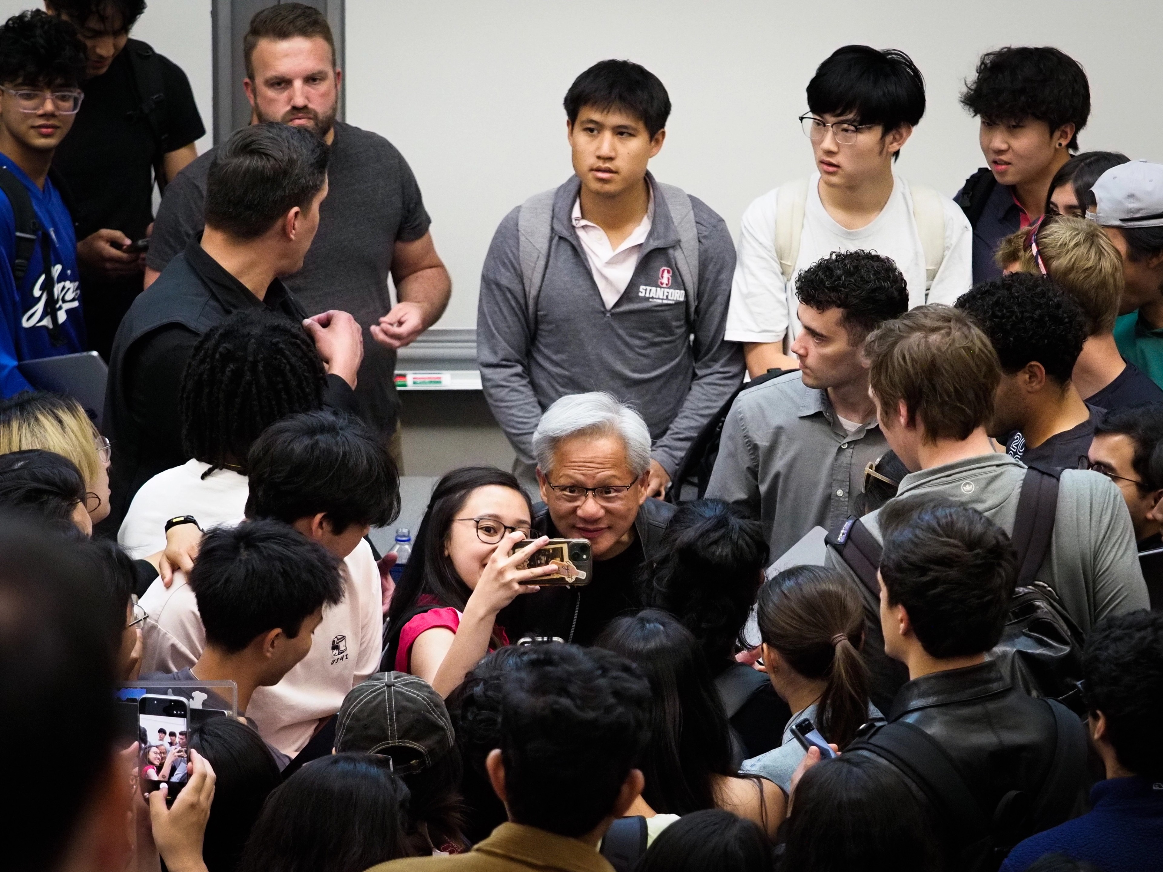 Jensen Huang at the center of a tight cluster of Stanford students, a young woman in a white shirt and red scarf in the foreground looking up at him smiling, a phone visible upper right.