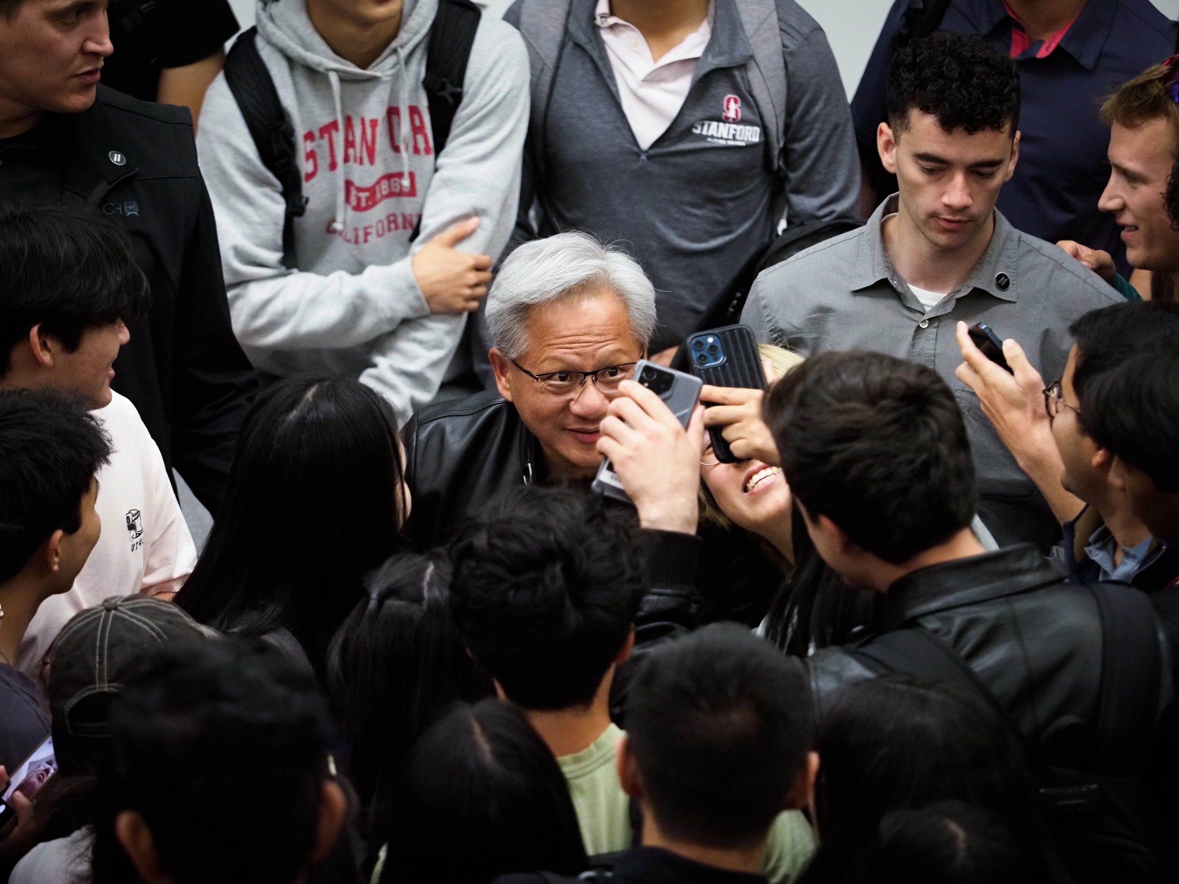 Jensen Huang surrounded by Stanford students, looking down at a phone screen one of them is holding up to him, another student in a Stanford-California sweatshirt visible in the crowd.