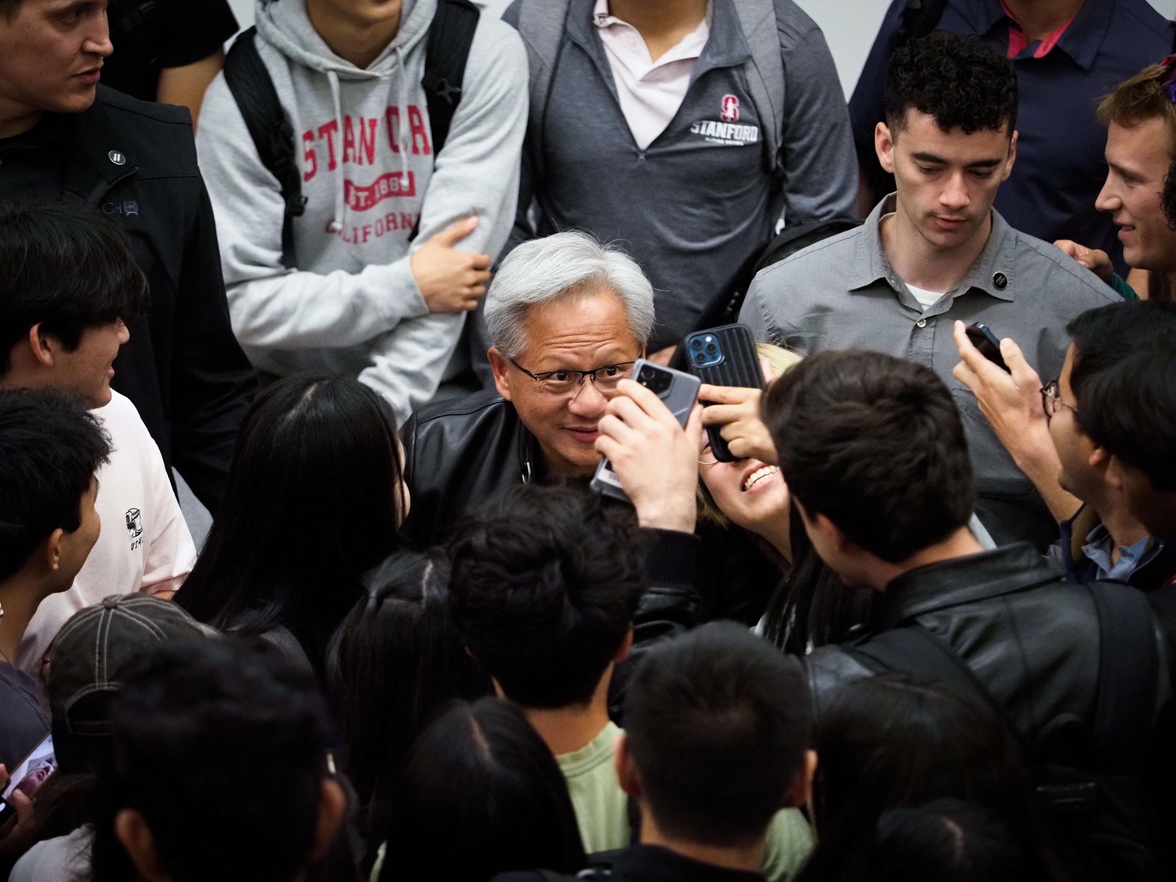 Jensen Huang surrounded by Stanford students, looking down at a phone screen one of them is holding up to him, another student in a Stanford-California sweatshirt visible in the crowd.