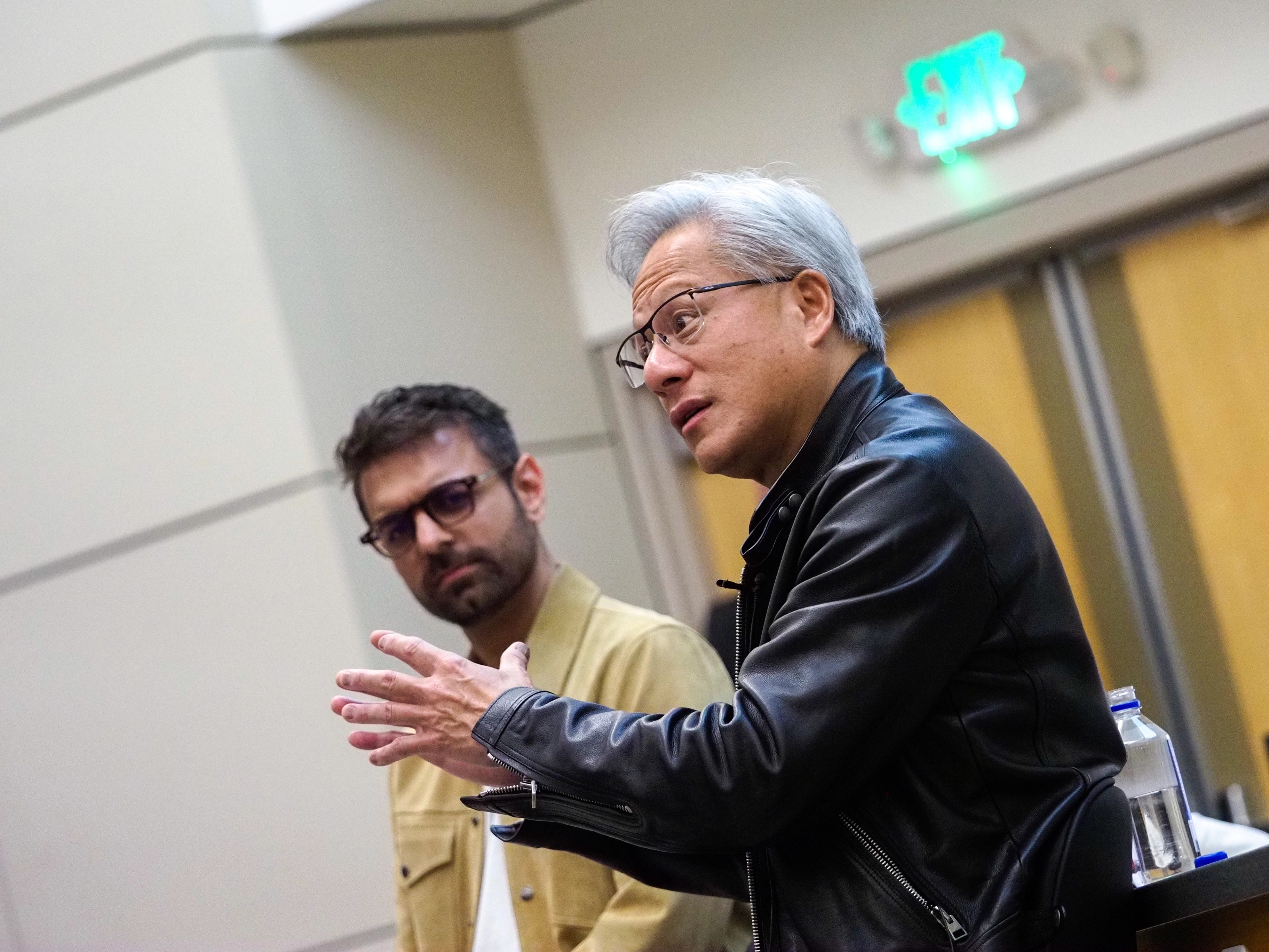 Jensen Huang in a black leather jacket gestures with one hand mid-thought as a bearded man in glasses and a mustard corduroy jacket listens beside him, both seated in a Stanford classroom.