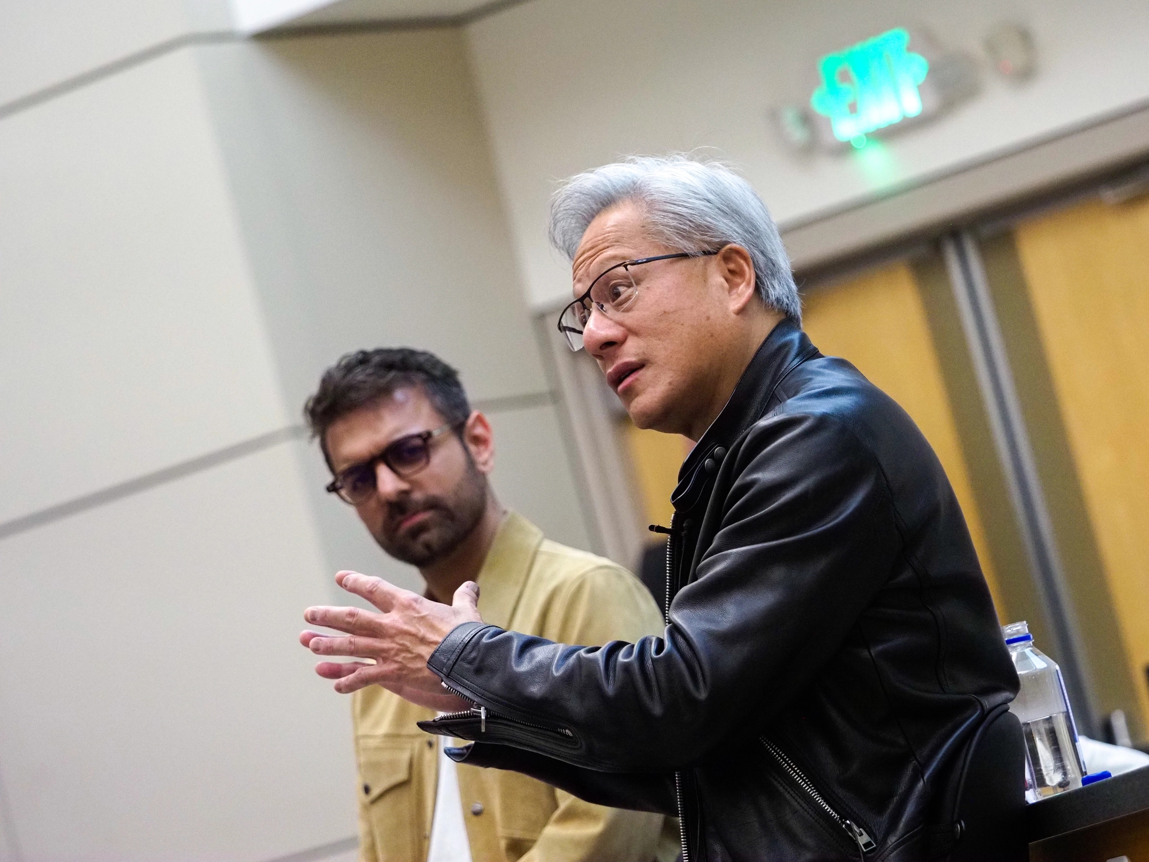 Jensen Huang in a black leather jacket gestures with one hand mid-thought as a bearded man in glasses and a mustard corduroy jacket listens beside him, both seated in a Stanford classroom.