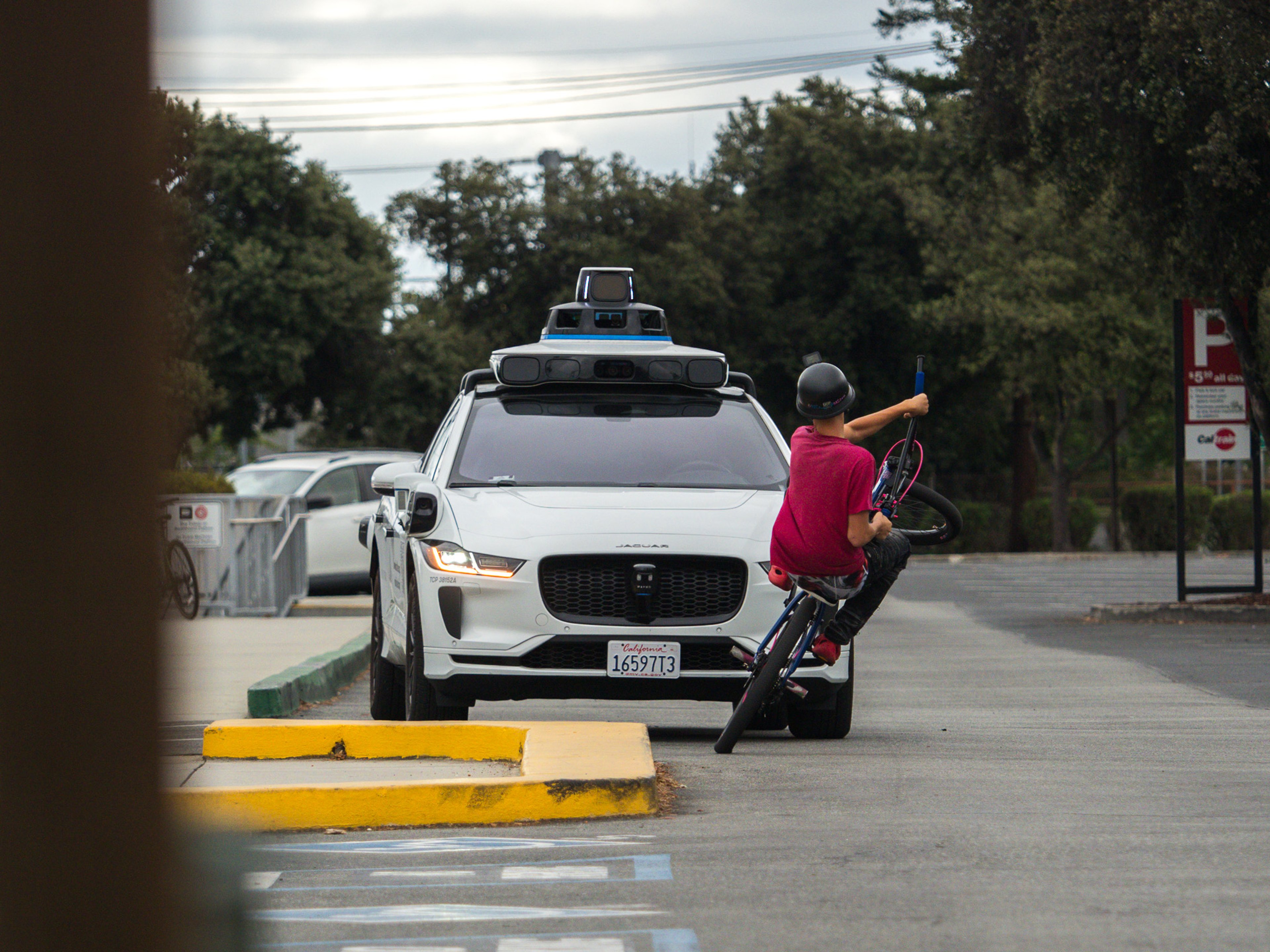 A young man on a BMX bicycle pulls a wheelie in a parking lot directly in front of an approaching white Waymo robotaxi with rooftop lidar sensors, looking back over his shoulder.