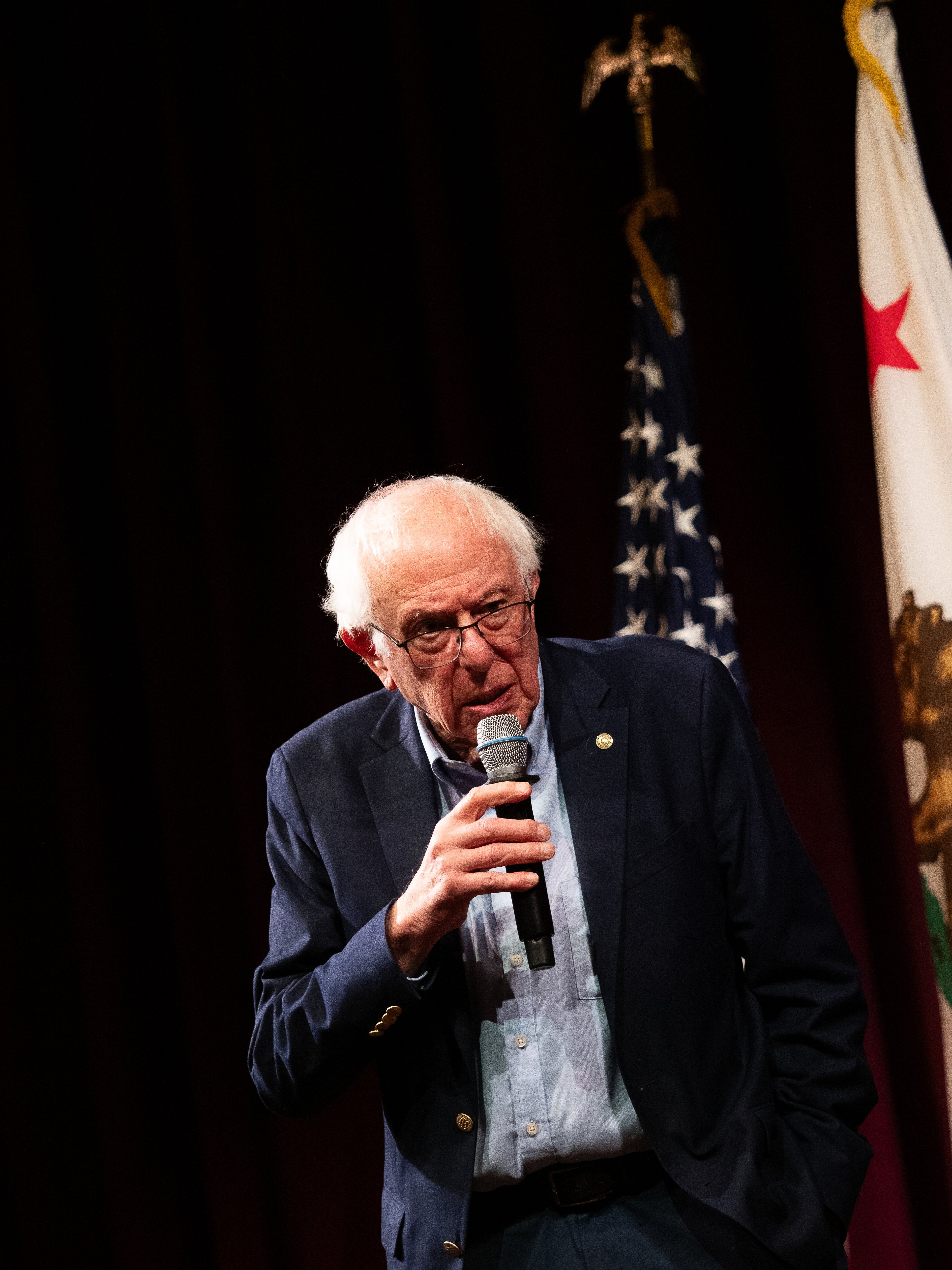 Bernie Sanders speaking with palm raised mid-gesture, microphone at chest height.
