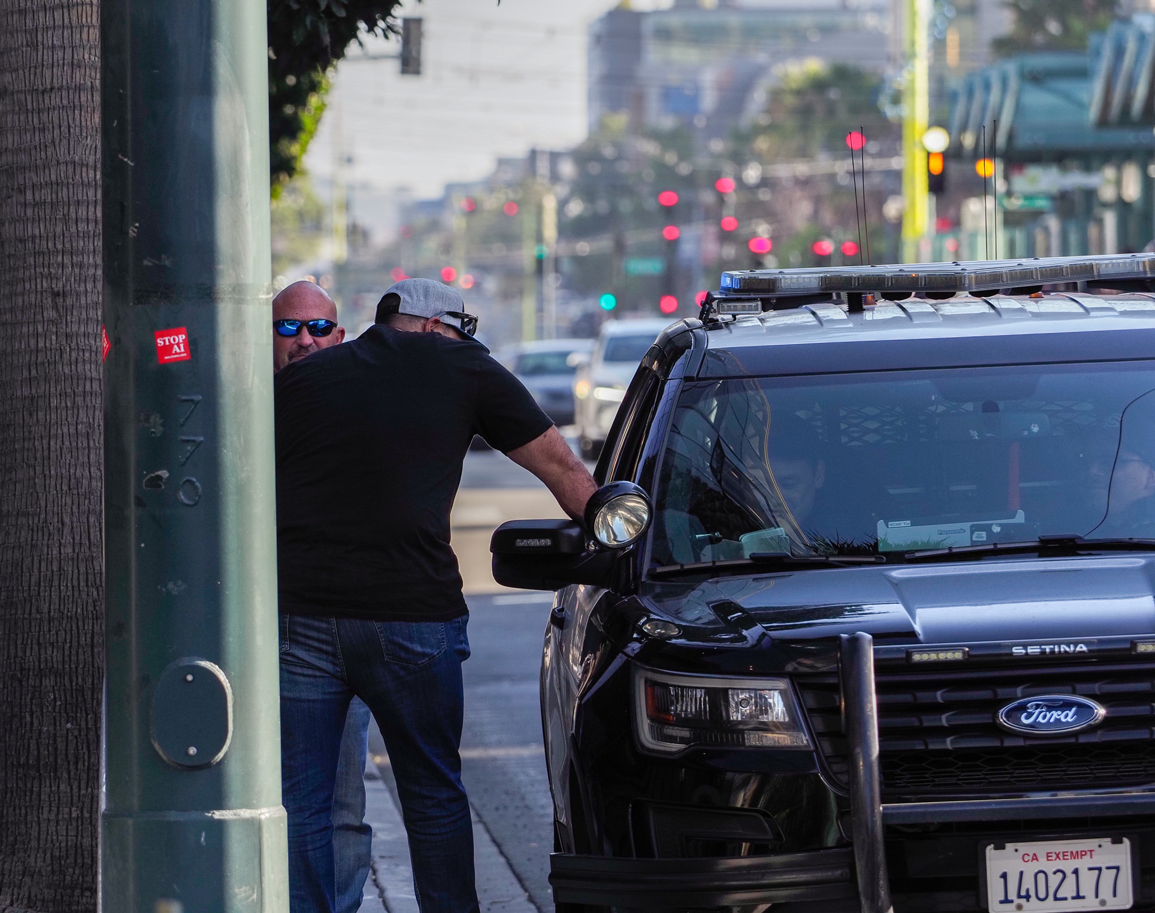 A bald man in a black t-shirt and jeans leans into the open passenger window of a black Ford SUV marked with police lights, framed by a green street pole on a downtown SF street.
