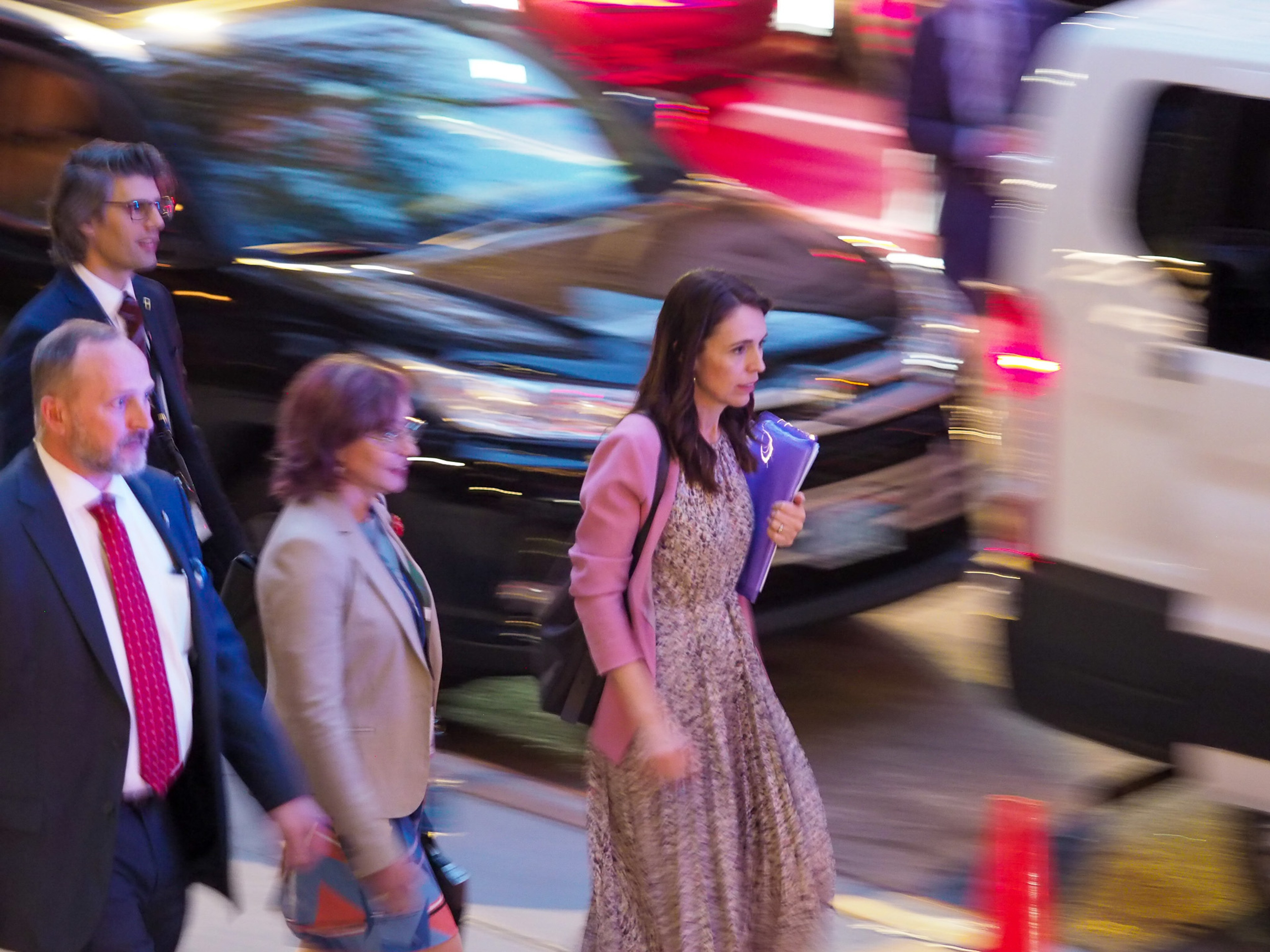 New Zealand Prime Minister Jacinda Ardern in a pink blazer and patterned dress walks past a passing motorcade rendered as streaks of red and white light by a slow shutter, two aides flanking her, a navy folder in her hand.