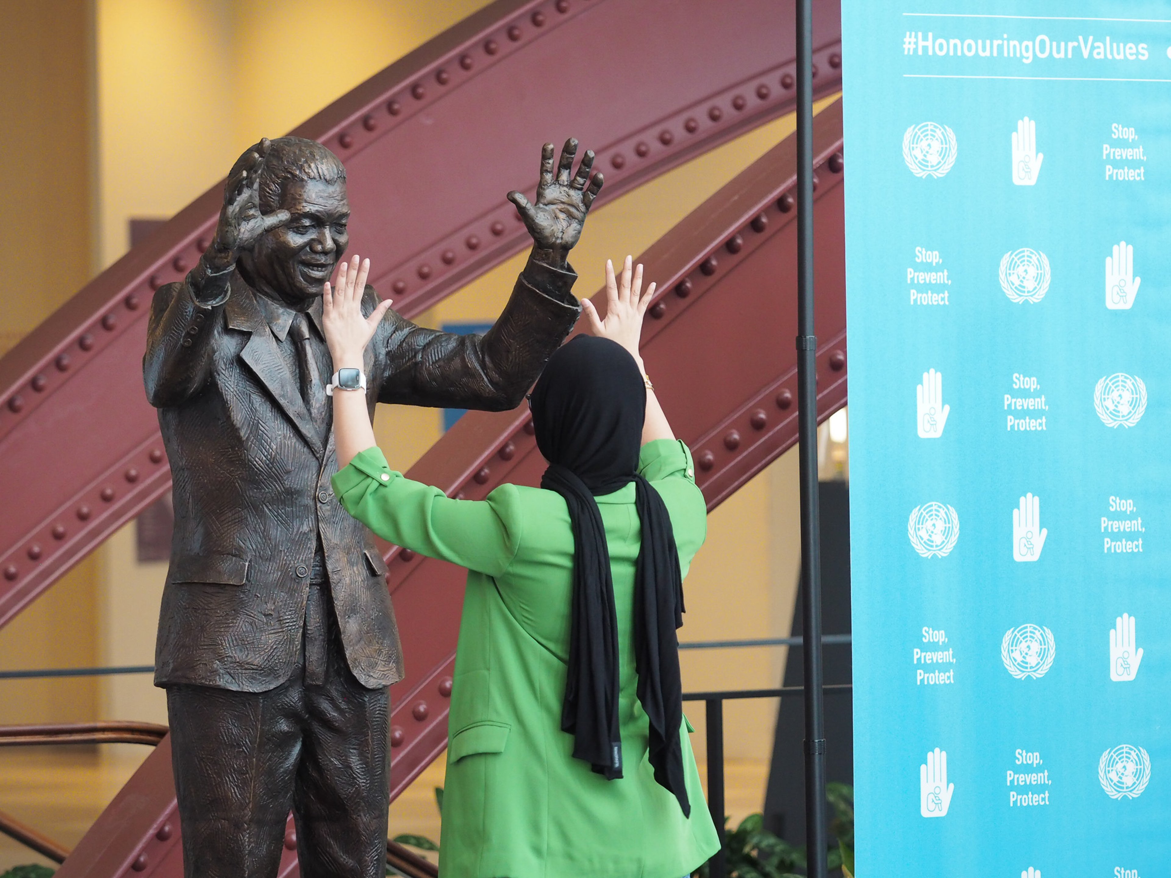 A woman in a green blazer and black hijab, photographed from behind, raises both her hands toward the upraised hands of a life-size bronze statue of Nelson Mandela inside the United Nations headquarters, beside a blue UN banner reading 'Honouring Our Values' and 'Stop, Prevent, Protect'.