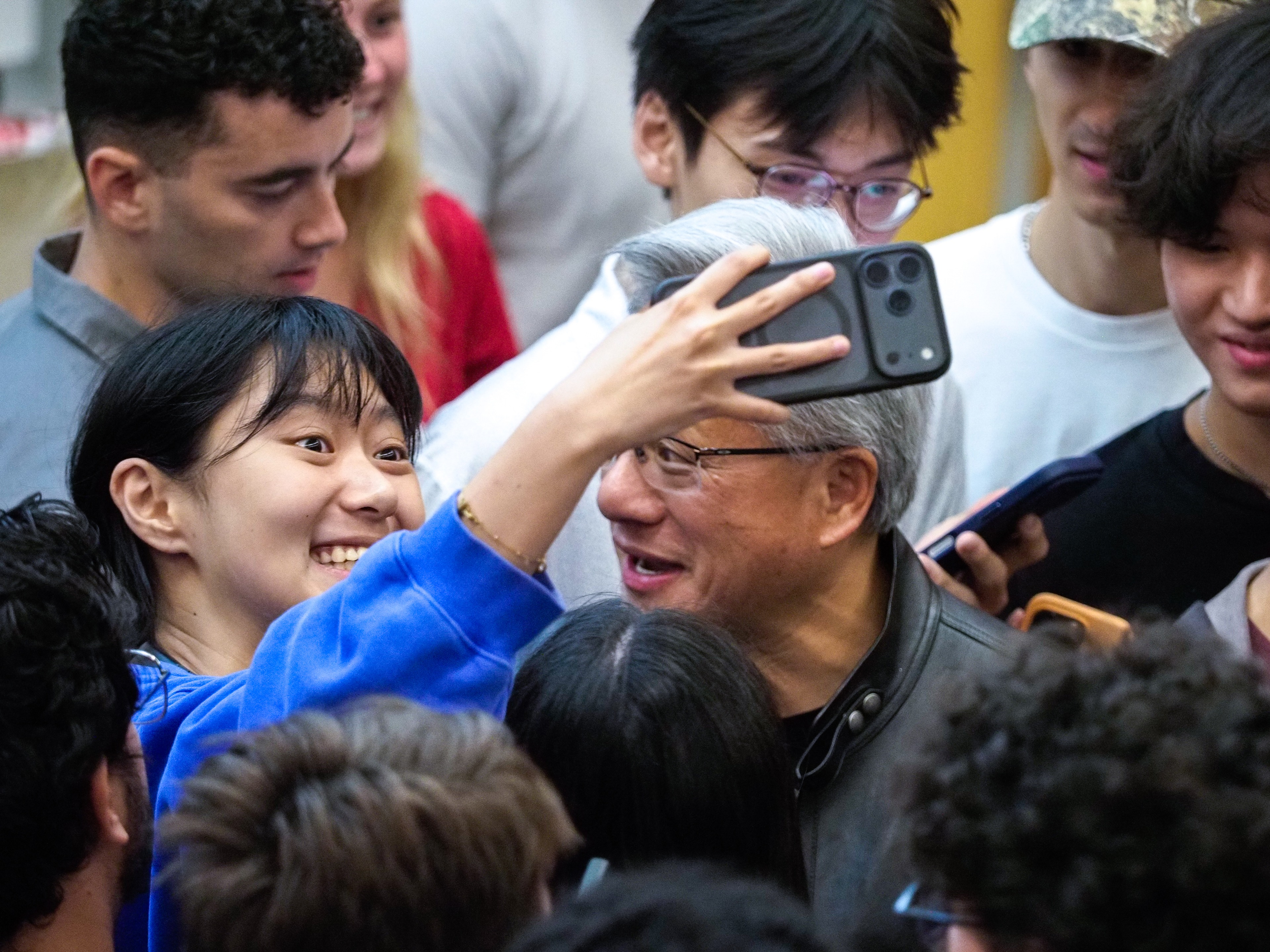 A young woman in a blue sweater holds up an iPhone for a selfie with Jensen Huang, both leaning in close, surrounded by other students reaching with their own phones.
