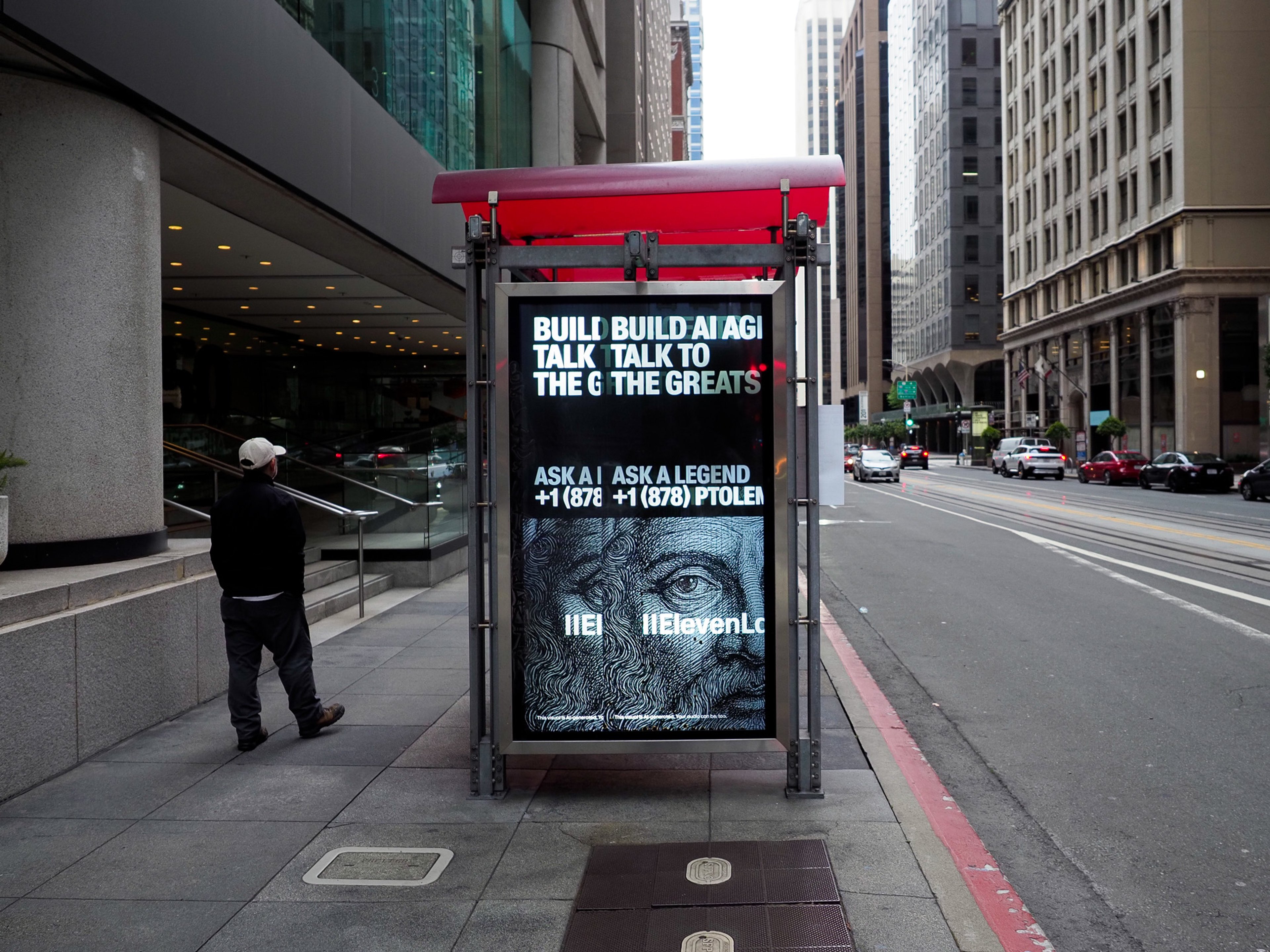 A pedestrian in a white cap walks past a downtown San Francisco bus shelter where two electronic poster panels mid-transition spell 'BUILD BUILD AI AGI' and 'TALK TALK TO THE GREATS', advertising ElevenLabs.