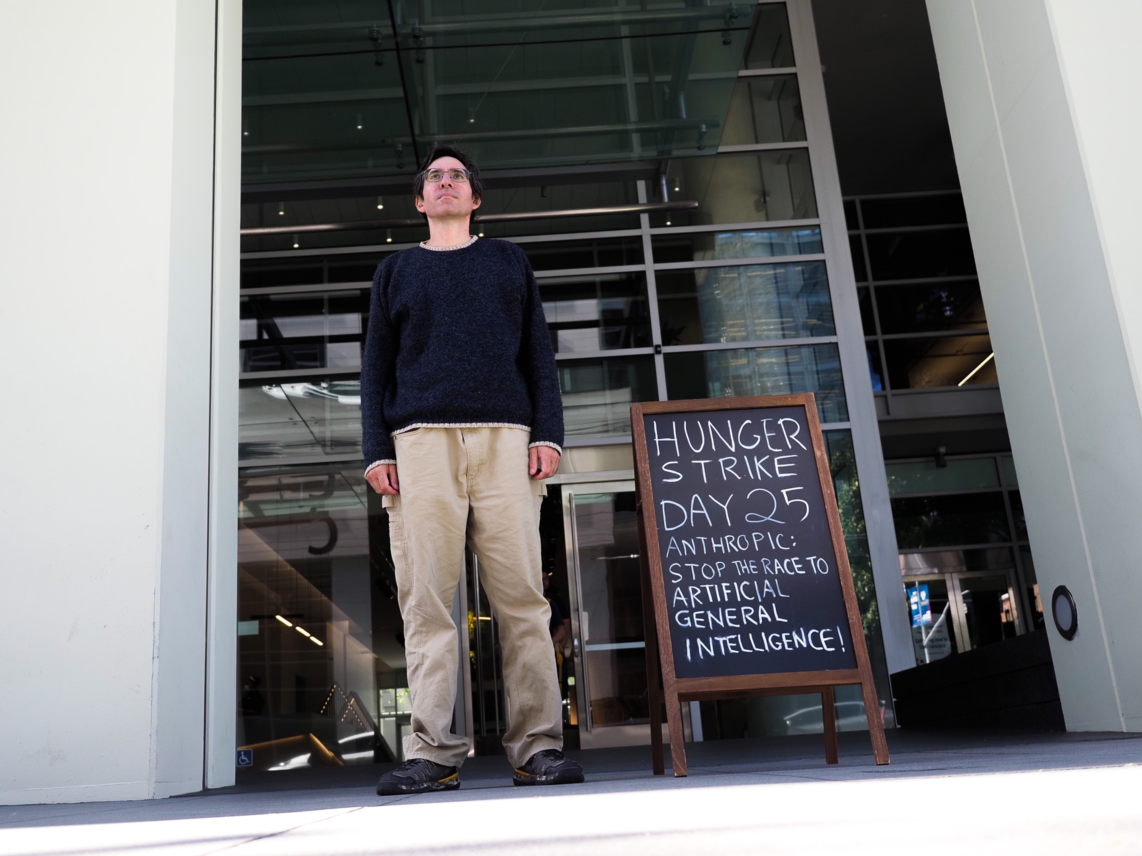 A man stands beside a chalkboard sign reading 'HUNGER STRIKE DAY 25 ANTHROPIC: STOP THE RACE TO ARTIFICIAL GENERAL INTELLIGENCE' outside a glass-fronted office building.
