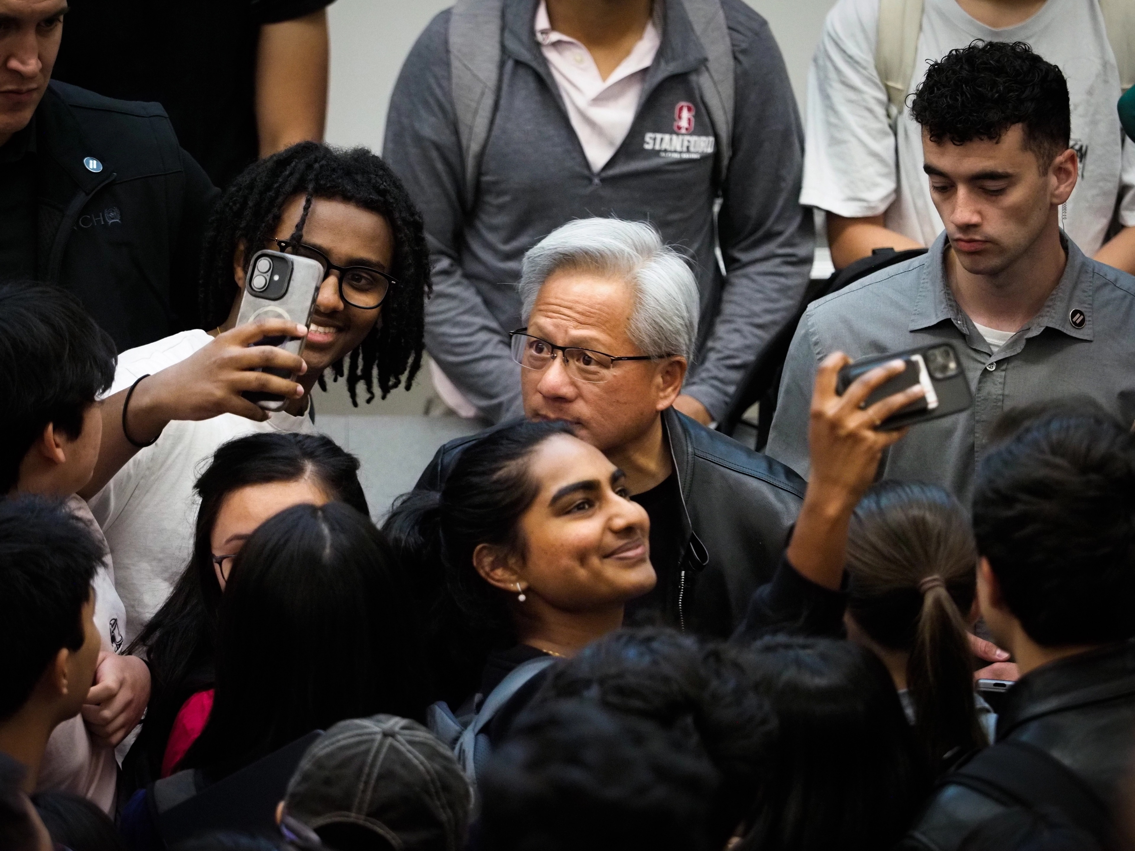 Tight crop of Jensen Huang's face mid-conversation, surrounded by Stanford students in sweatshirts, partially obscured by an outstretched arm and the back of a student's head.