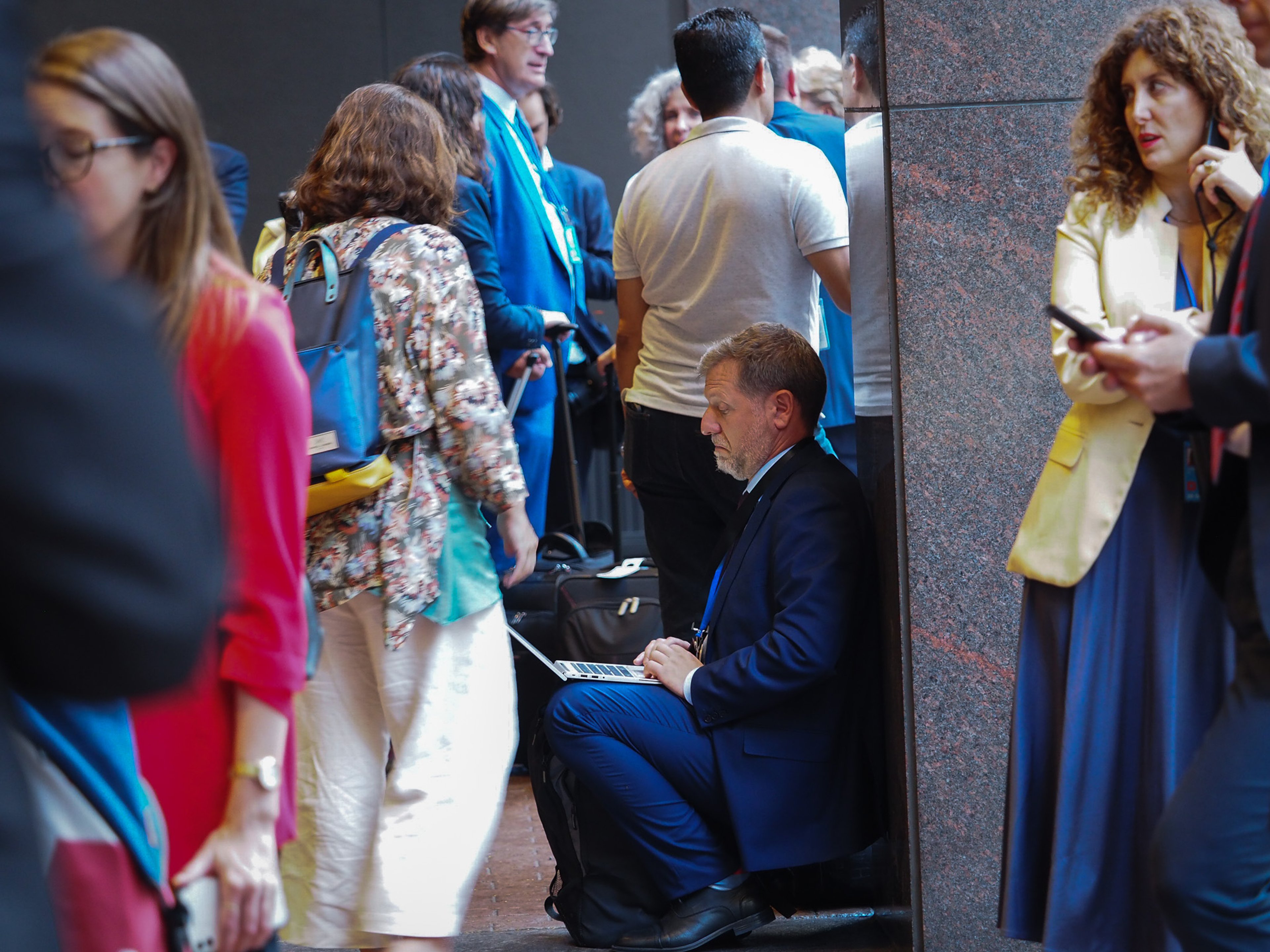 A man in a dark suit crouched on the floor against a black marble wall, working on a laptop balanced on his lap, surrounded by delegates and journalists in motion.