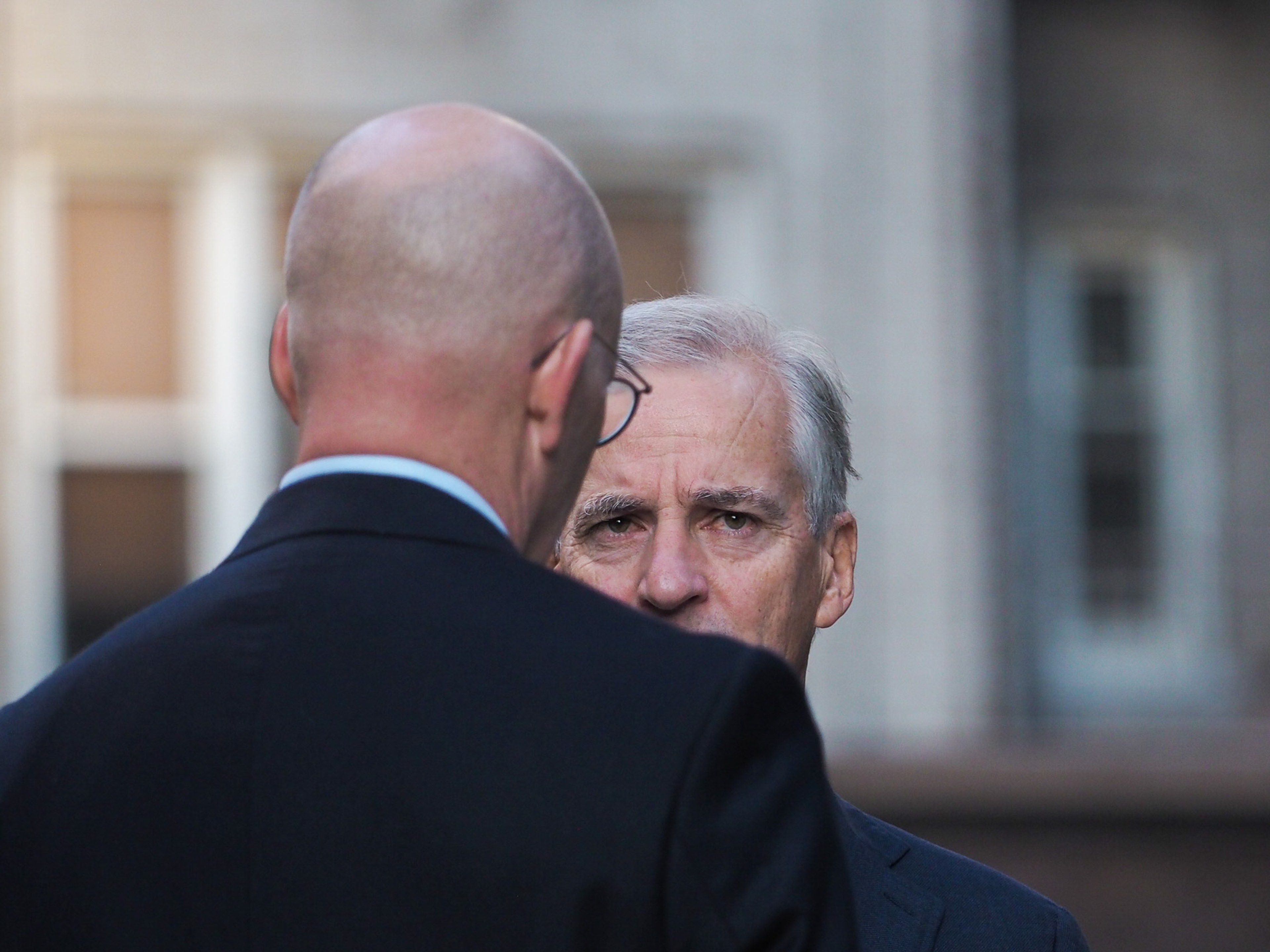 Tight portrait of Norwegian Prime Minister Jonas Gahr Støre in profile listening, the back of an interviewer's bald head and shoulder filling the foreground left of frame.
