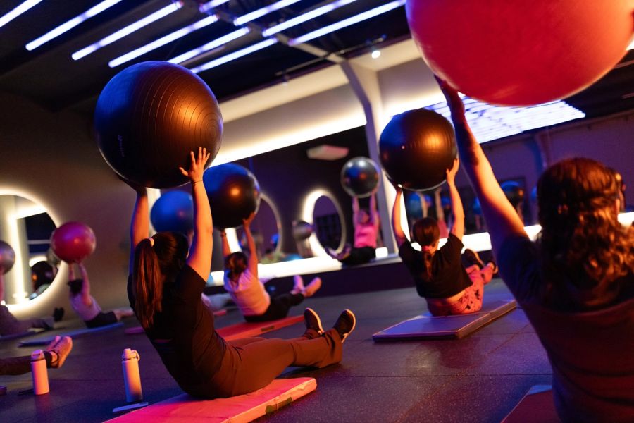 Alumnas de Pilates sentadas en la colchoneta sosteniendo el balón sobre la cabeza