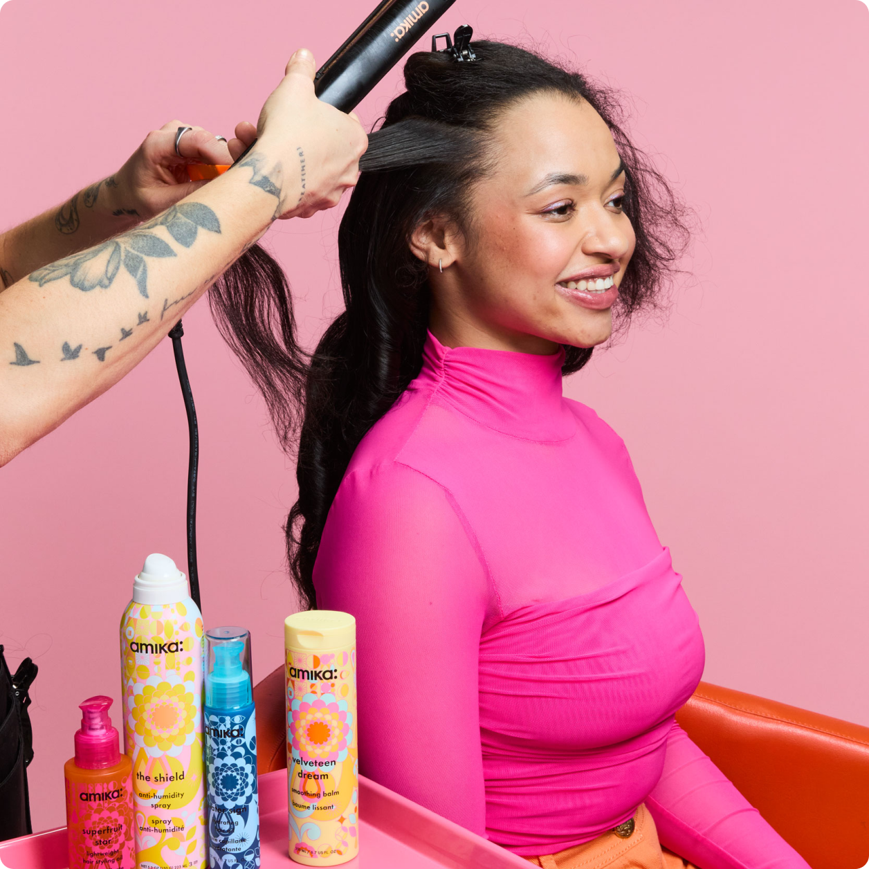 a stylist holds a flat iron near a model's hair. there are amika products on a stand next to the chair.