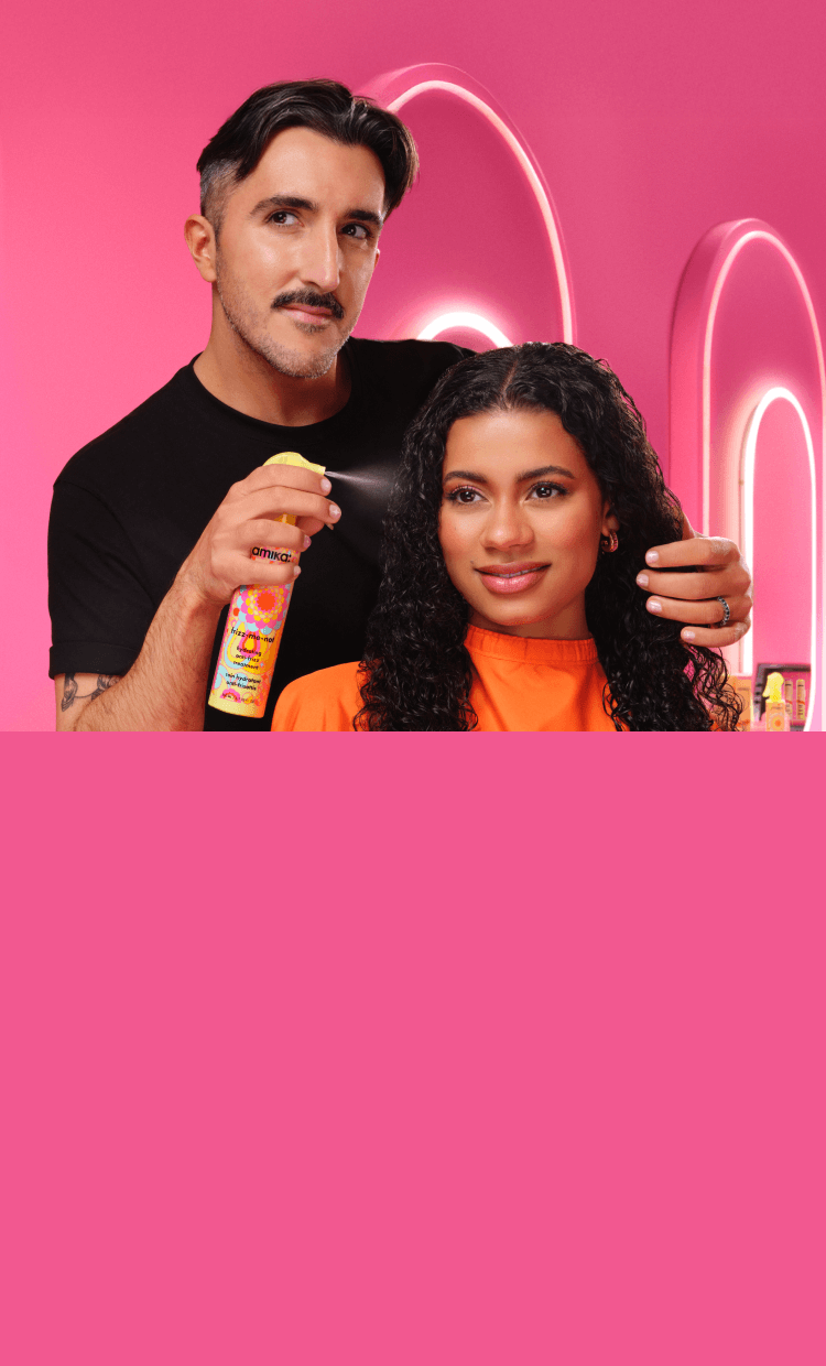 a stylist applies amika's frizz-me-not hydrating anti-frizz treatment spray to a person with curly hair in a pink salon environment