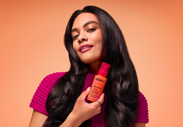 a model holds a bottle of superfruit star hair oil against an orange background