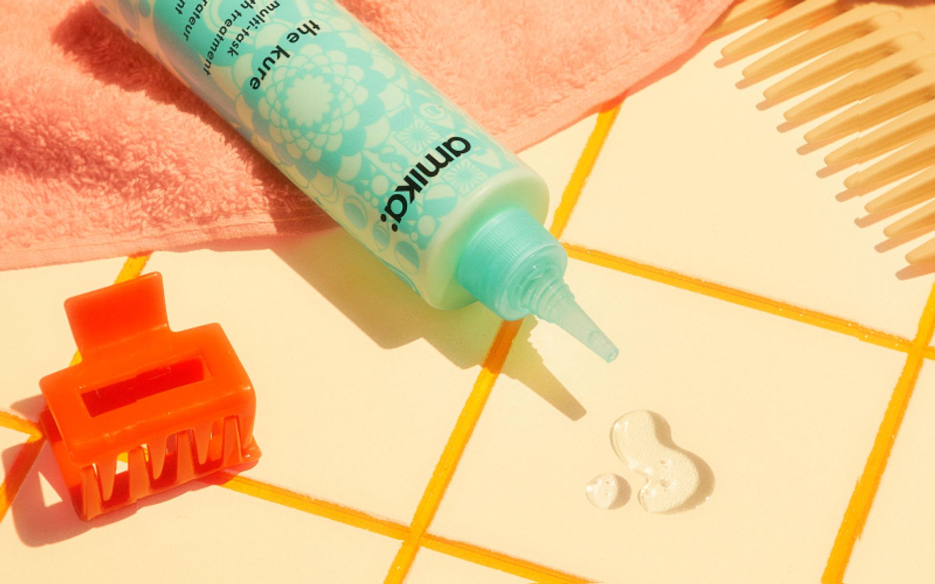 bottle of amika the kure multitask strength repair treatment slightly spilling onto a yellow tiled surface with an orange clip and yellow comb nearby