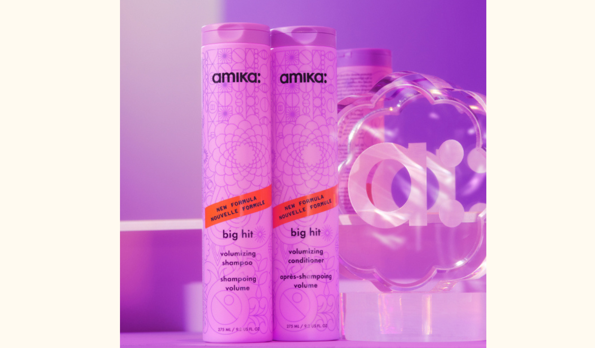 image of a close-up display of Amika's "Big Hit" volumizing shampoo and conditioner bottles set against a purple background. The two tall, decorative bottles are prominently positioned side by side, showcasing their intricate patterns and vibrant packaging. Behind the bottles, a clear decorative piece with the Amika logo is visible, adding to the brand's playful and stylish aesthetic.