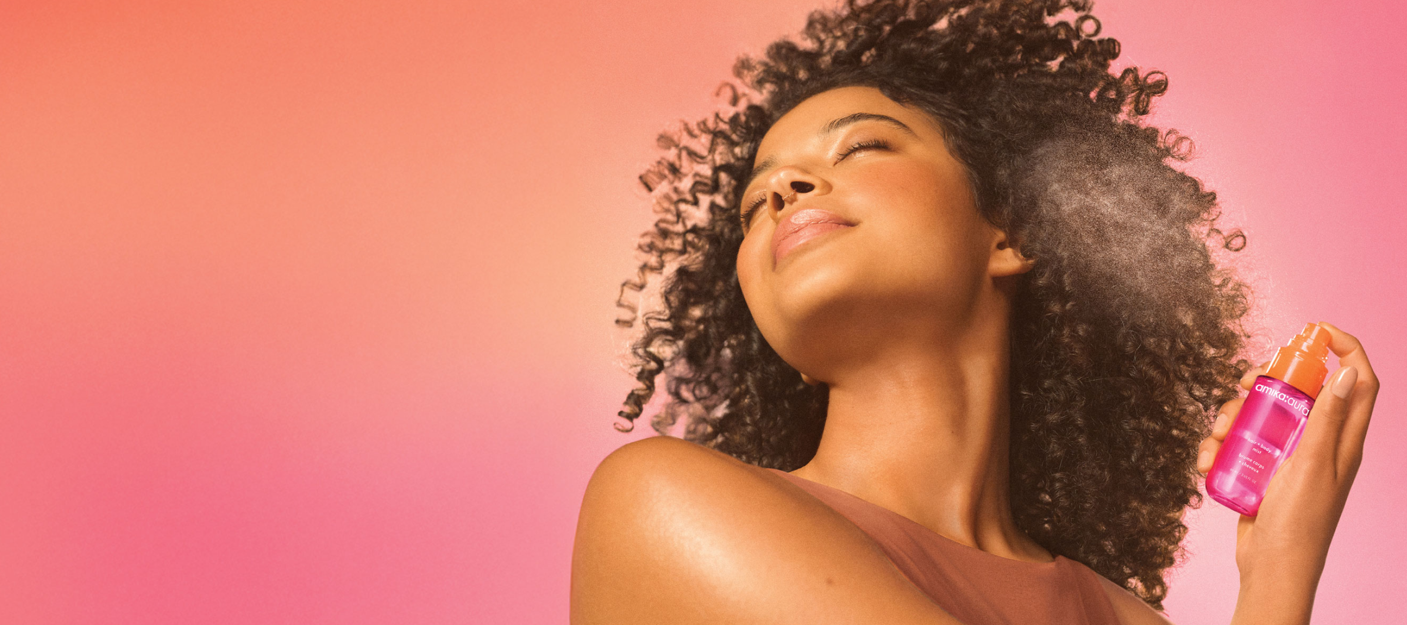 a model mists amika:aura hair and body mist onto their hair. model is set against a pink gradient background