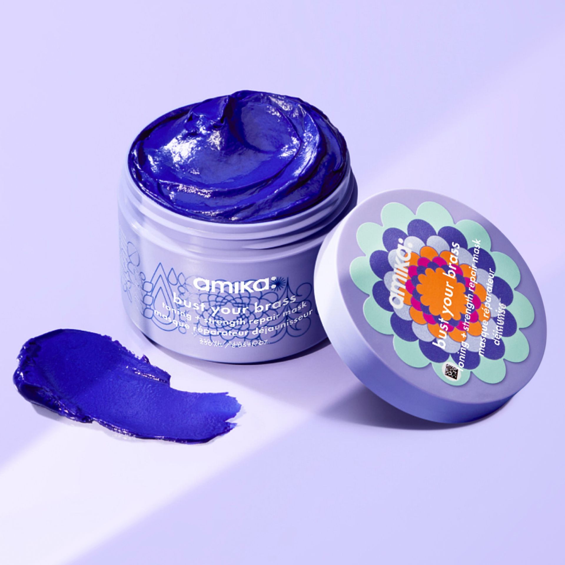 image of a jar of amika bust your brass intense strengthening repair mask. the lid is leaned against the canister with the purple mask product exposed and a smear of purple mask on the light purple background.