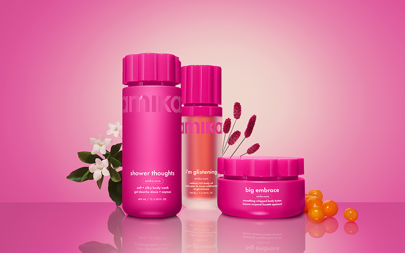 amika shower thoughts body wash, i'm glistening body oil, and big embrace body butter sit against a pink gradient background with flowers and sea buckthorn berries