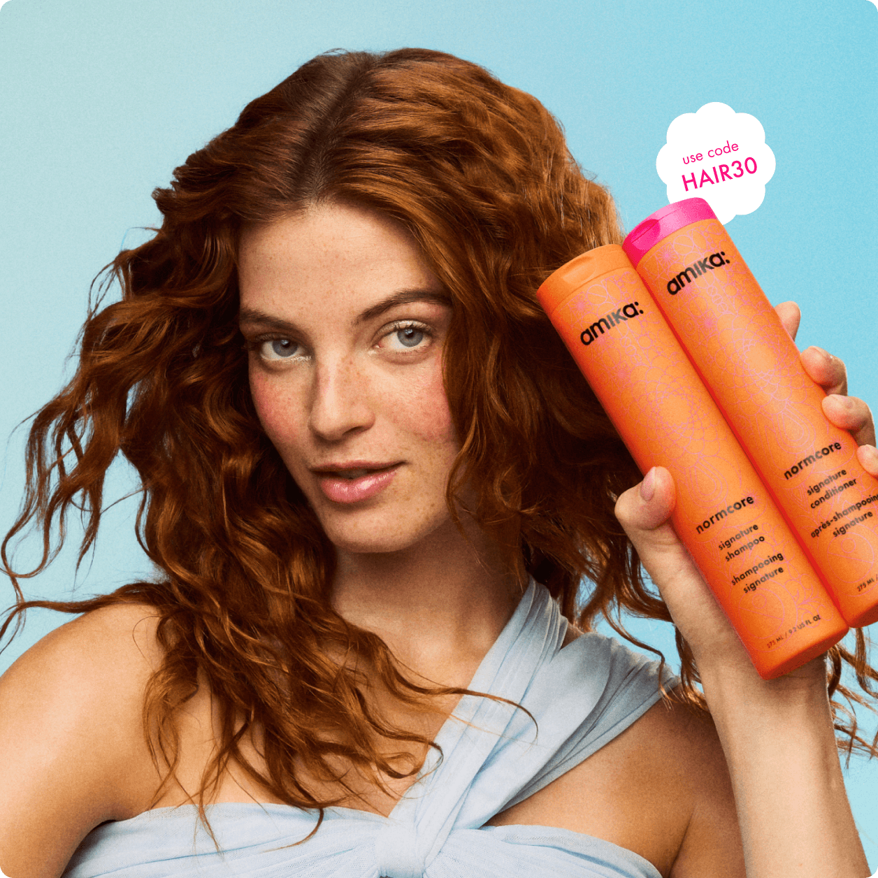 model with wavy red hair holds amika normcore shampoo + conditioner bottles against a light blue background