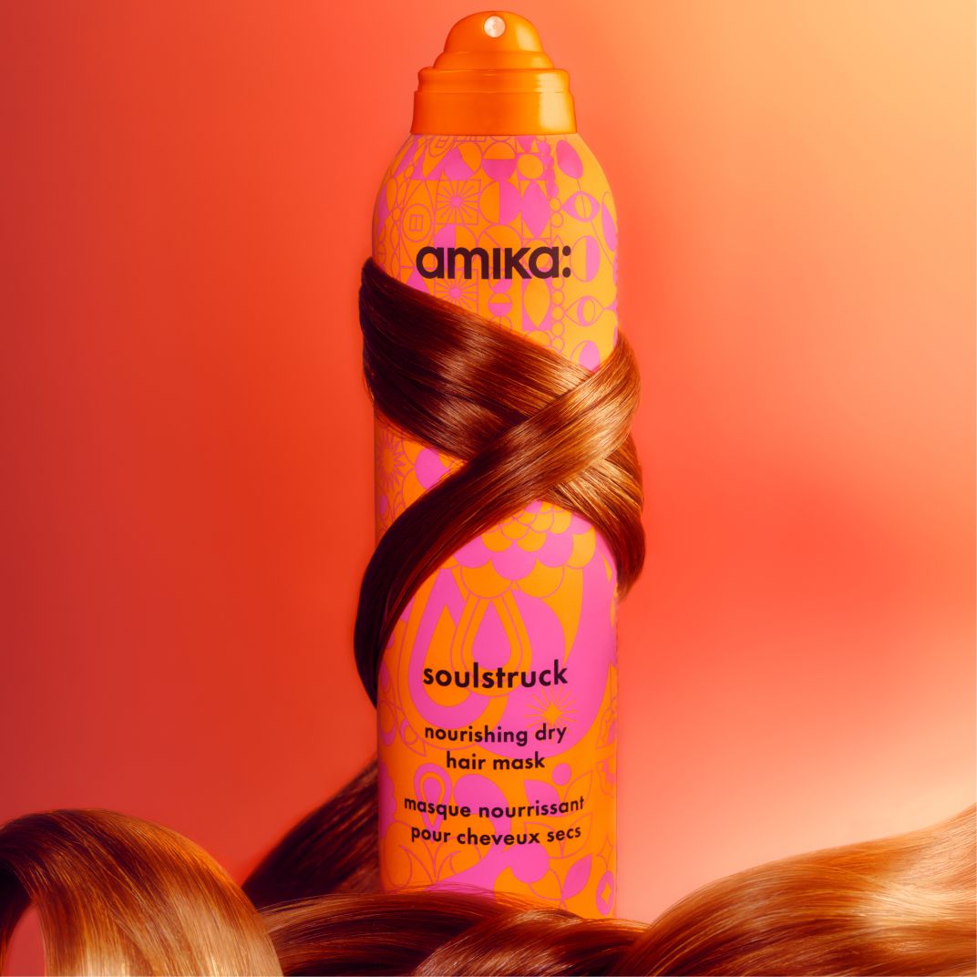 a tress of reddish brown hair wraps around amika soulstruck spray. the bottle sits in front of a pinkish-orange background