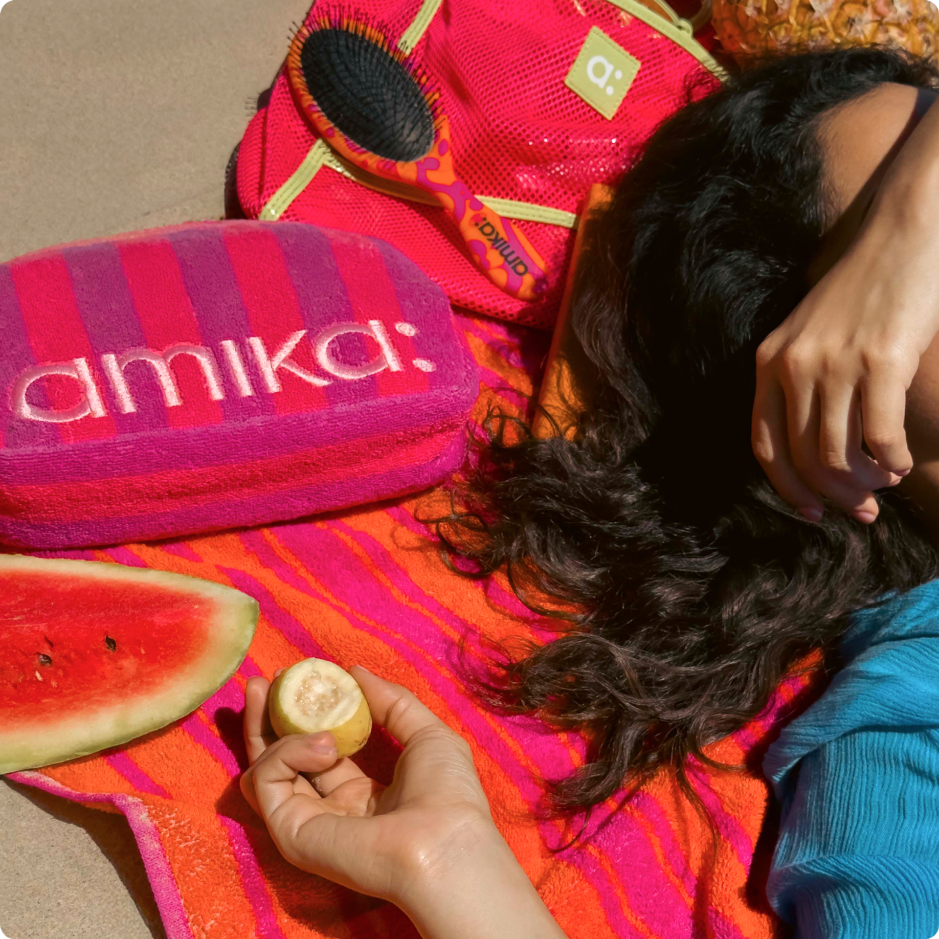 a model lies on a pink and orange towel with fruit and a pink and purple amika cosmetic bag and hairbrush nearby