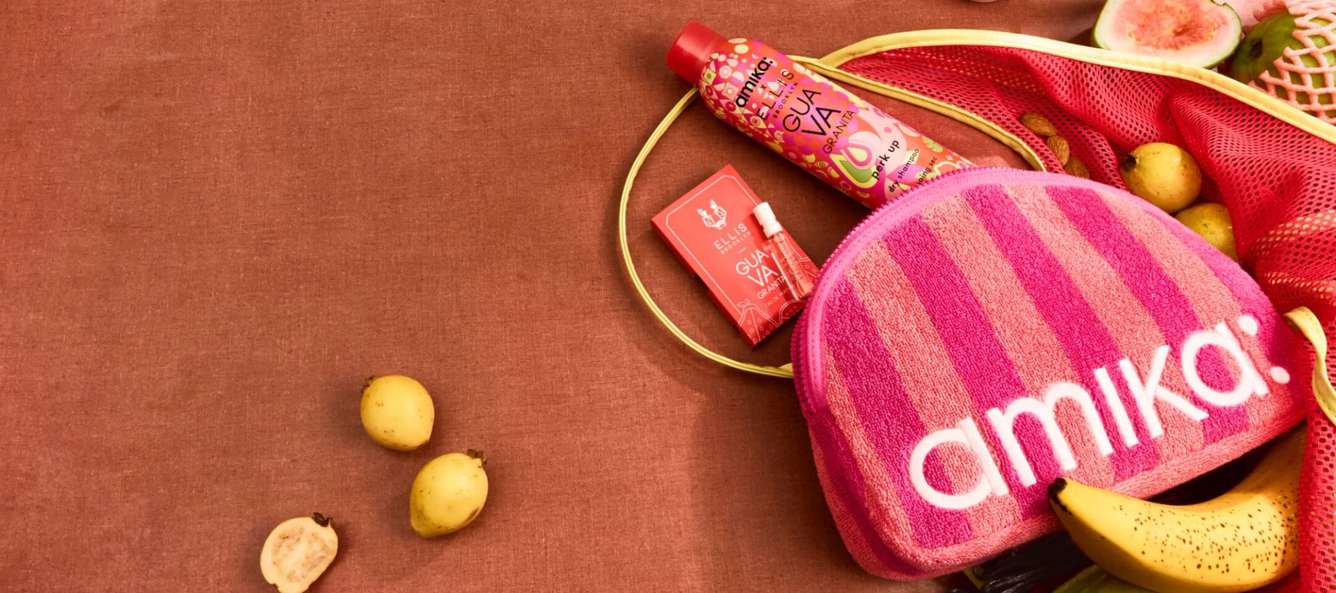 amika perk up GUAVA GRANITA, GUAVA GRANITA perfume sample, and pink striped logo terrycloth pouch lay amongst tropical fruits against a brown background