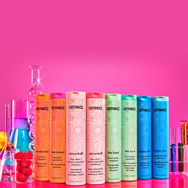 A bright and colorful display of Amika's hair care products featuring six bottles in different vibrant colors. Each bottle has a unique pattern and color, including the normcore shampoo and conditioner, mirror ball shampoo and conditioner, the kure shampoo and conditioner, the hydro rush shampoo and conditioner. The bottles are arranged in a row against a vivid pink background. Surrounding the bottles are various laboratory glassware, such as beakers and test tubes, filled with colorful liquids, along with fresh fruits and candy in glass jars, creating a lively and playful scene. The image emphasizes the energetic and fun branding of Amika products.